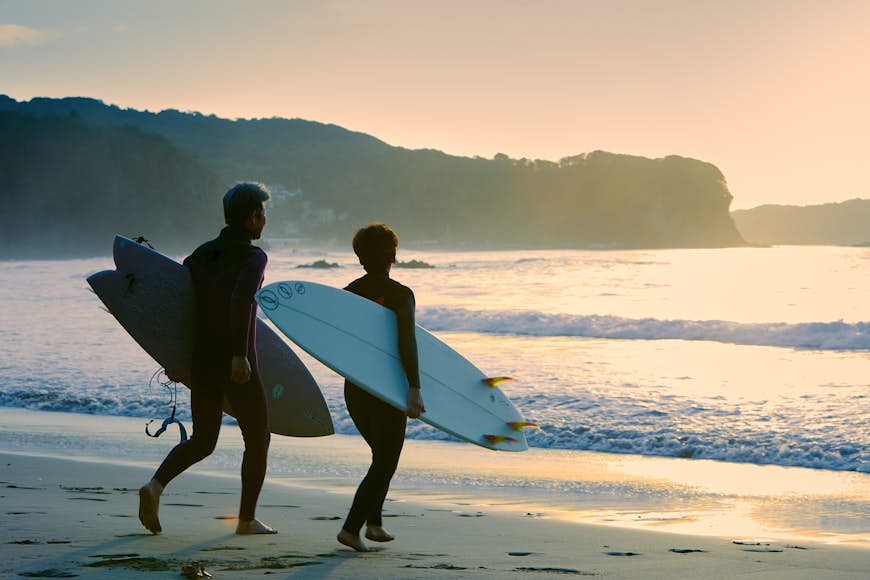A Japanese man and woman head into the sea in Shimoda with their surfboards under their arms. The sand beneath them is golden.
