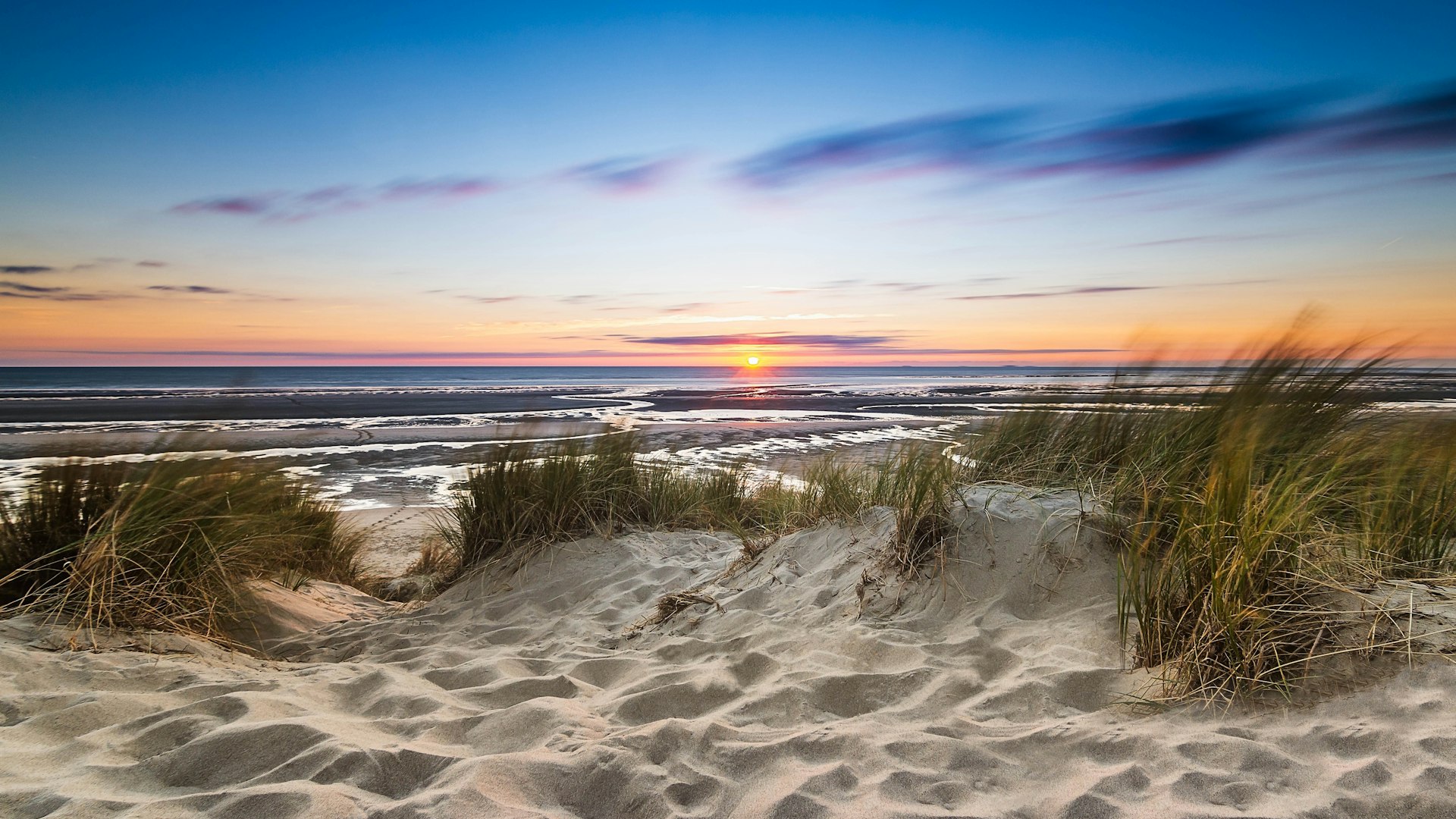 Sand dunes on the edge of a body of water with the sun setting 