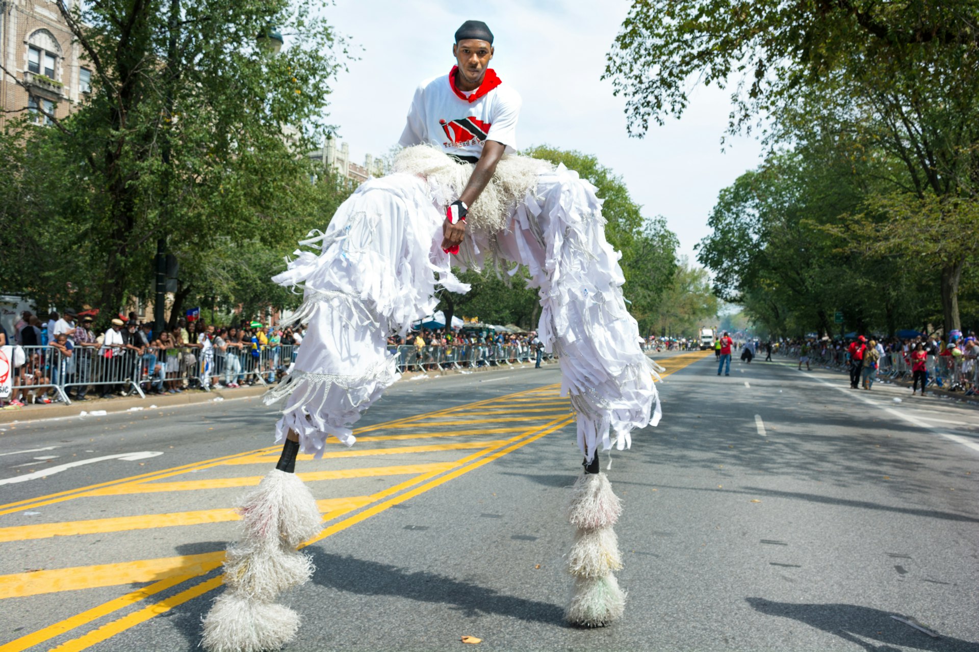 A man wearing stilts and wearing white tassel pants and a white t-shirt with the Trinidad and Tobago flag on it walks at West Indian Day Parade. 