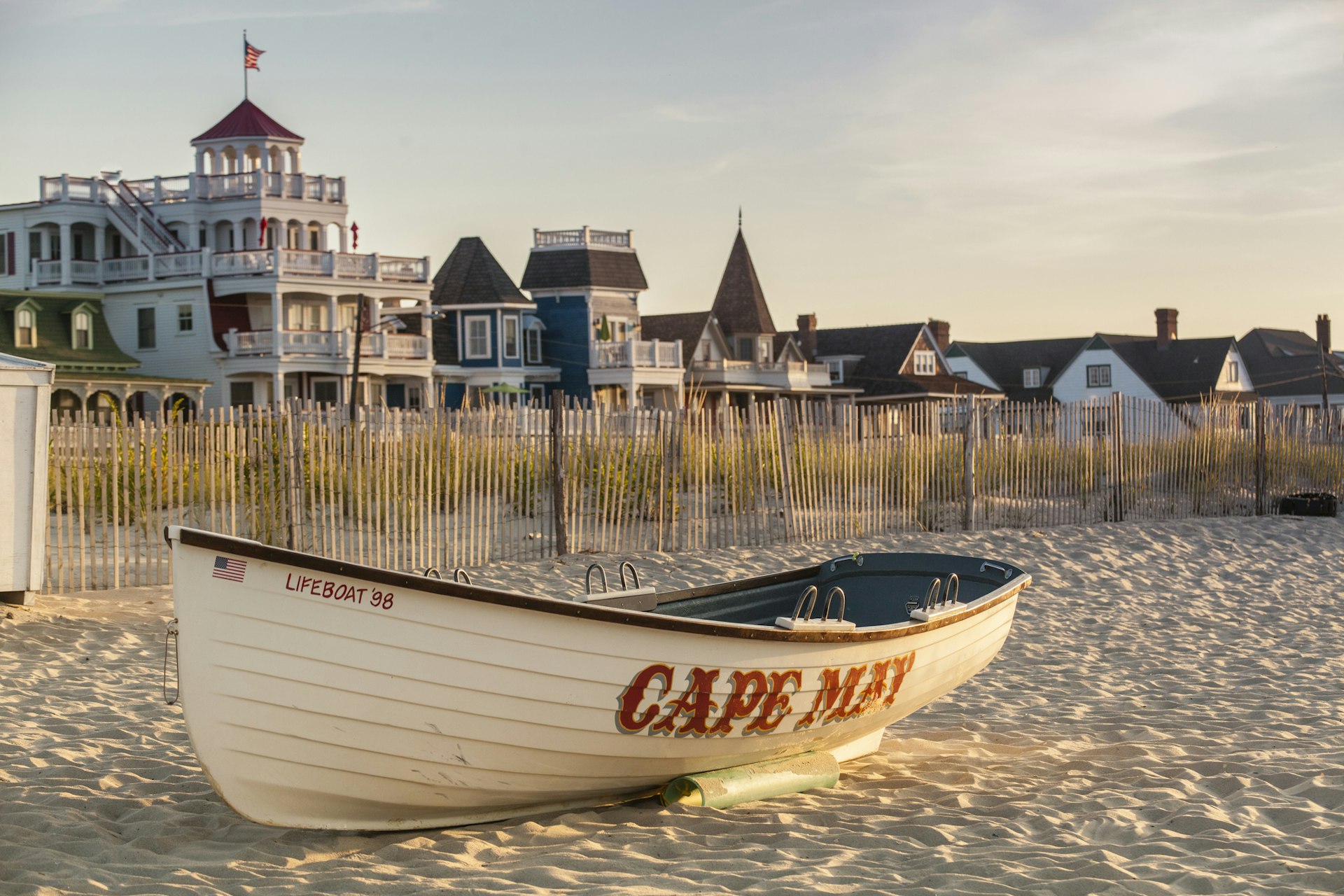 An empty wooded boat lays on the sand in Cape May. There are large Victorian Homes in the background. 