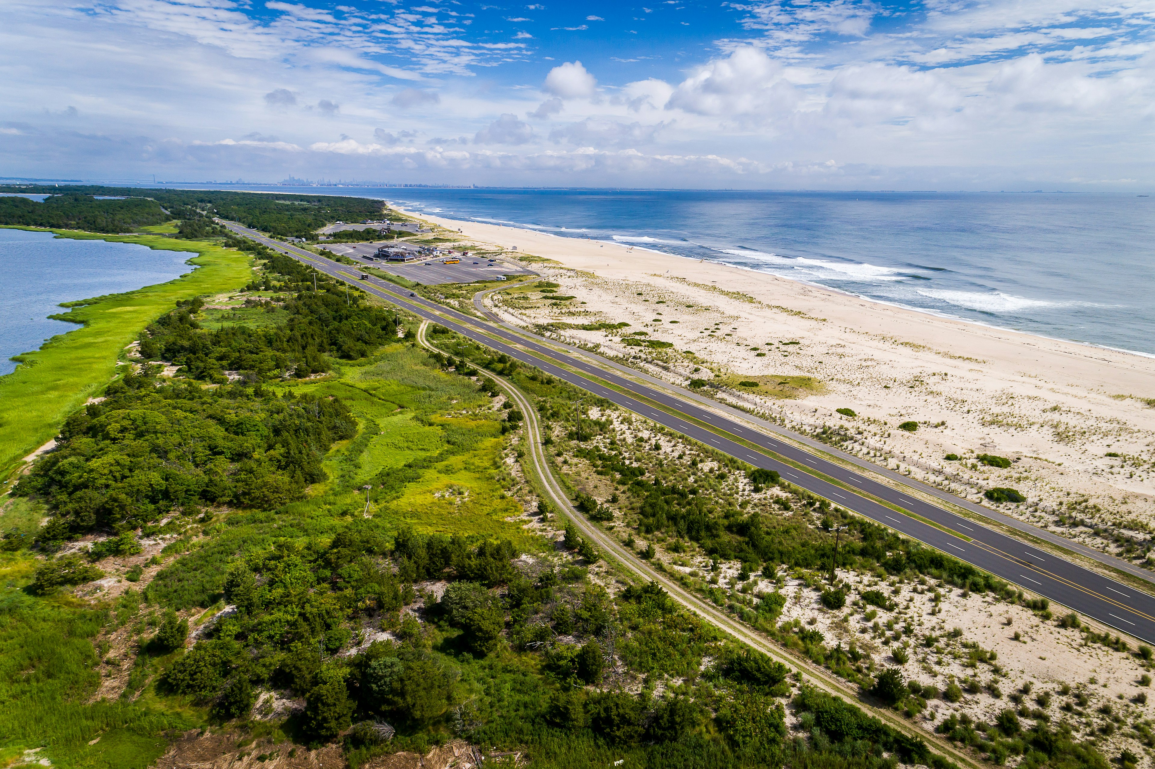 Aerial view of a peninsula with sandy beach on one side and greenery on the other. A road runs down the center