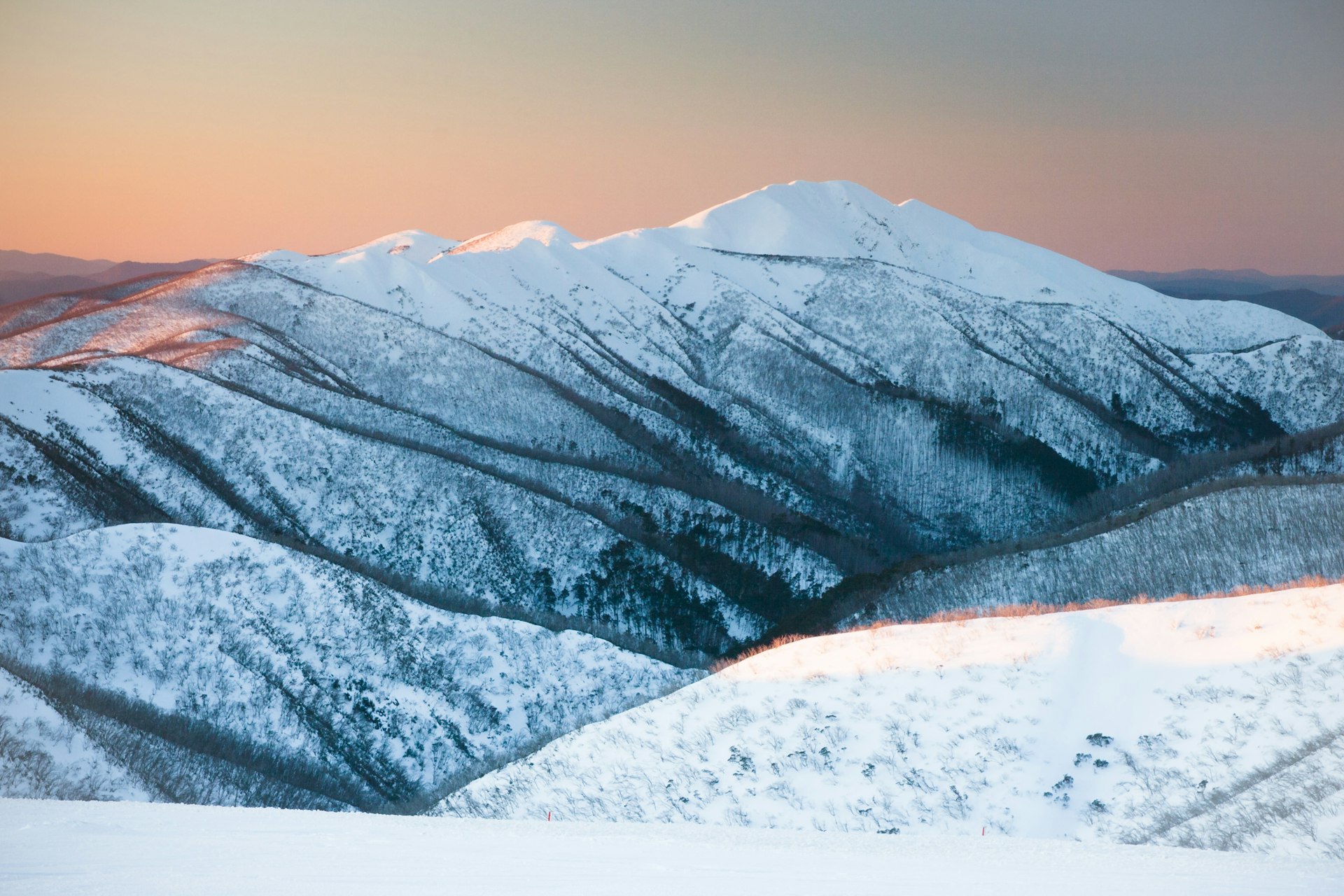 A blanket of snow sits on the rolling mountain range of Australia's Alpine National Park at sunset.