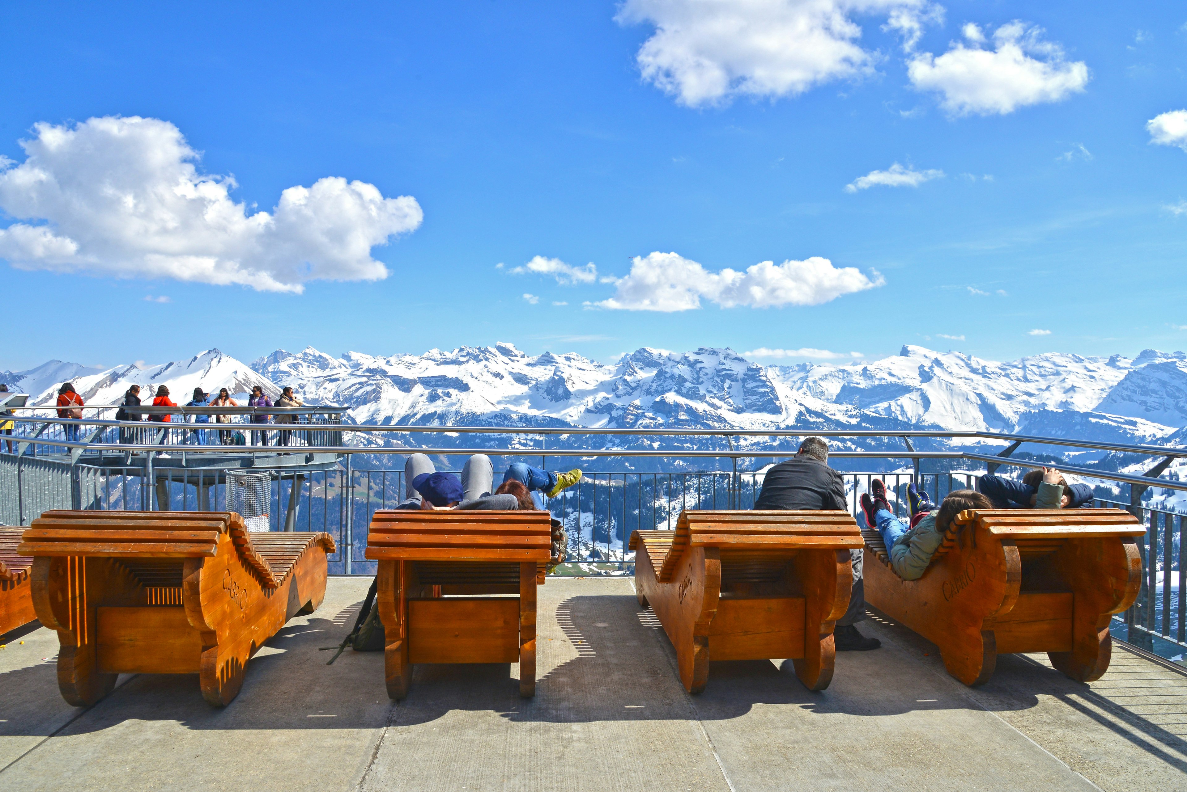 Visitors in wooden deck chairs above snow-capped mountains on the sun terrace atop Stanserhorn Mountain