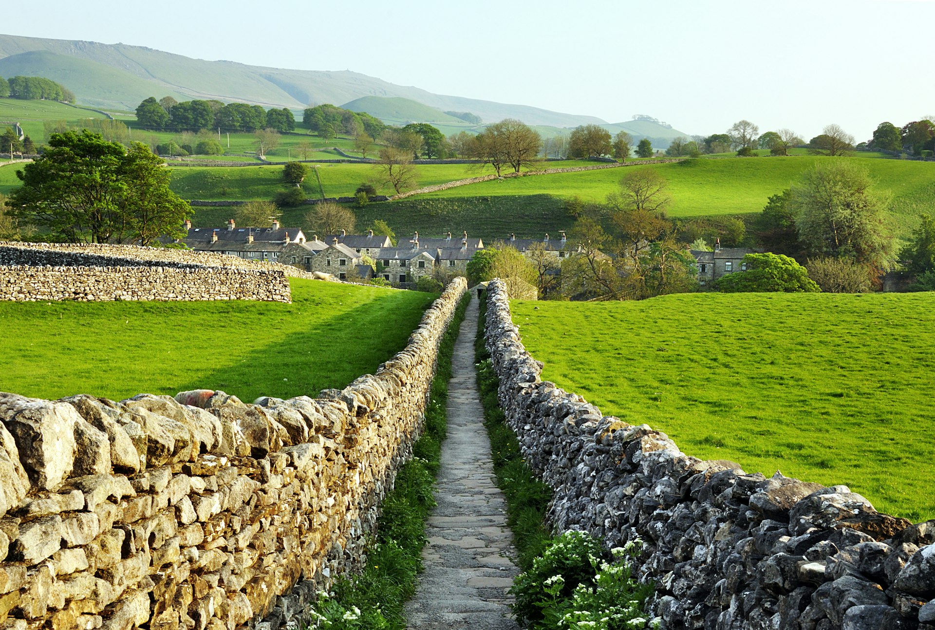 A path, lined with drystone walls, stretches out ahead through countryside leading to a village of stone houses. 