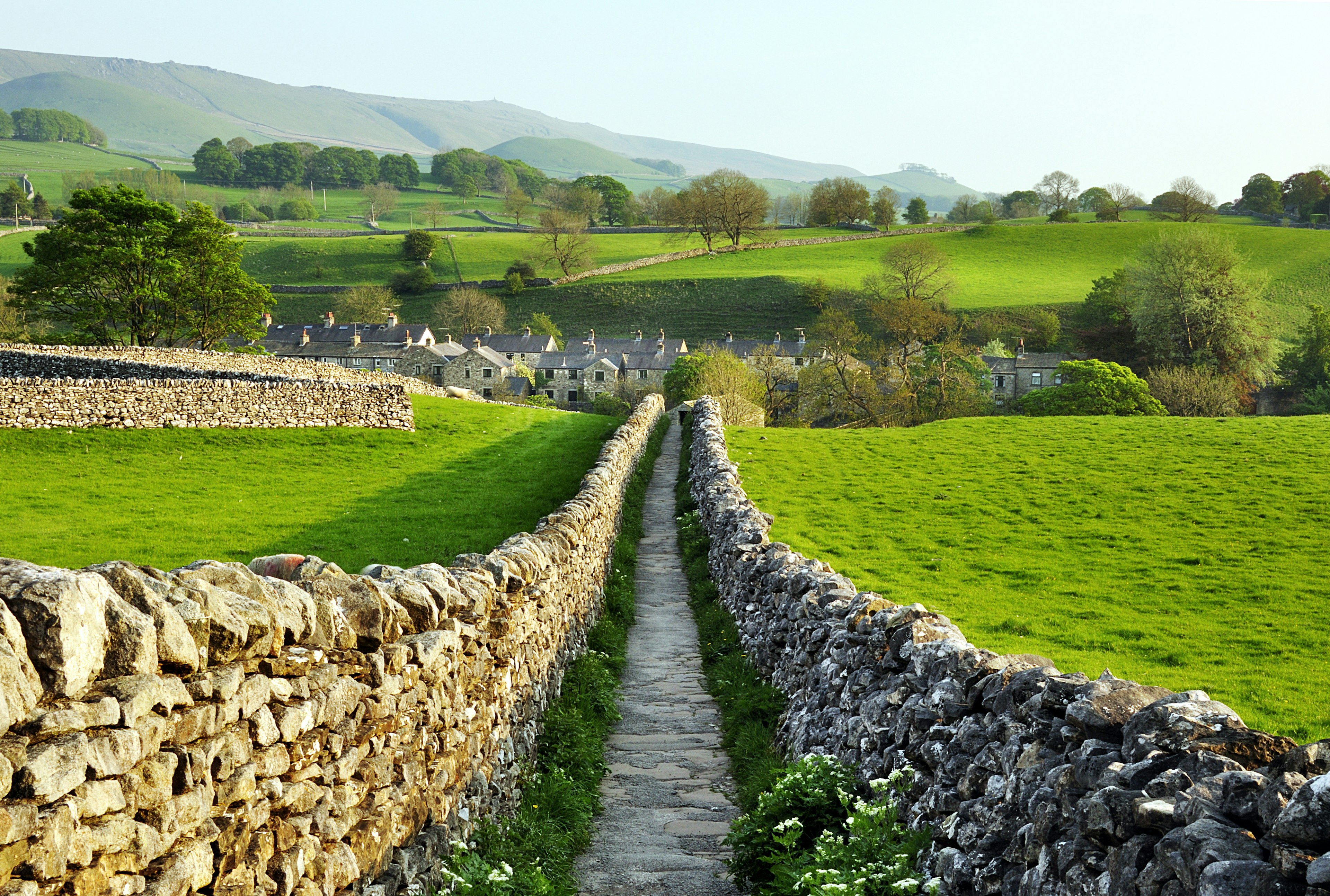 A path, lined with drystone walls, stretches out ahead through countryside leading to a village of stone houses.