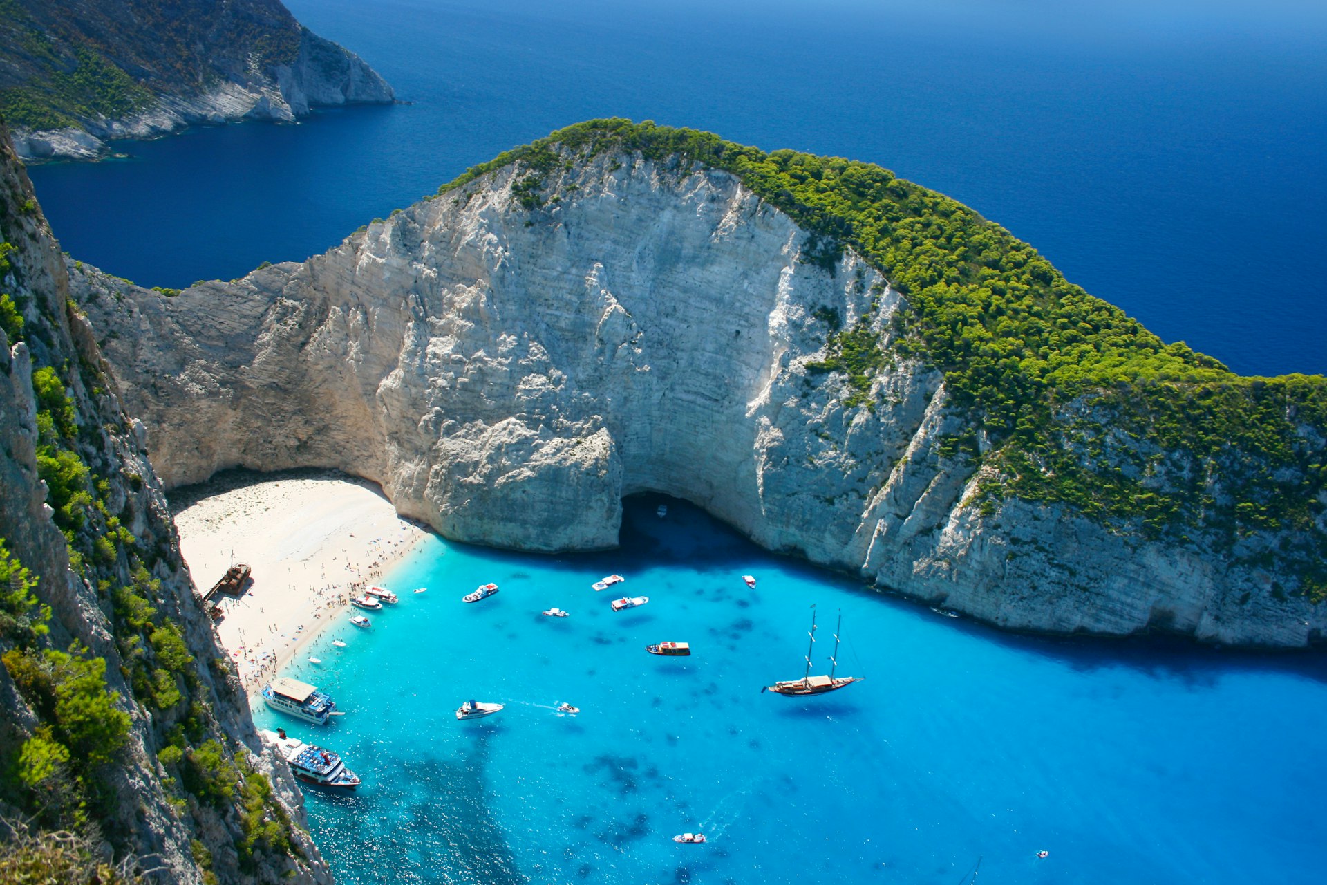 An aerial view of Navagio Beach, a sheltered cove in Greece with white sand, blue waters and a large wrecked ship on the shore.