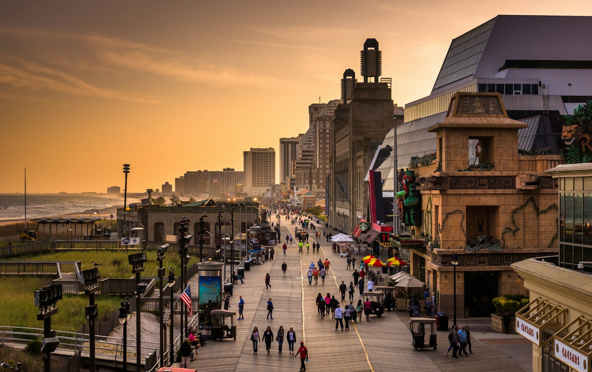 The sun is setting over a beach town. People are walking along the wide boardwalk lined with large buildings