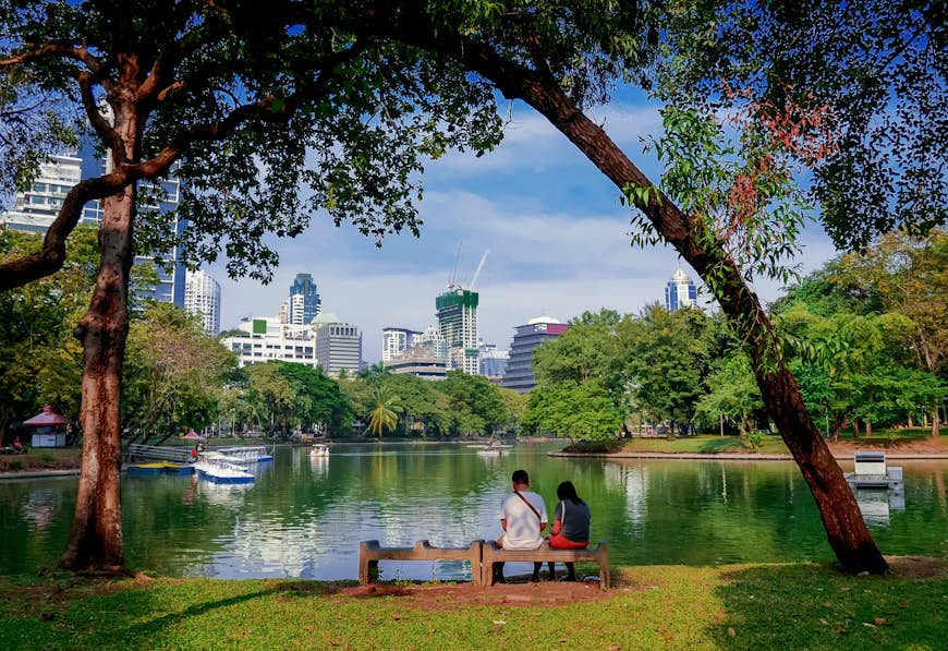 A couple sit next to one another on a bench which overlooks the lake at Lumpini Park in Bangkok, Thailand.