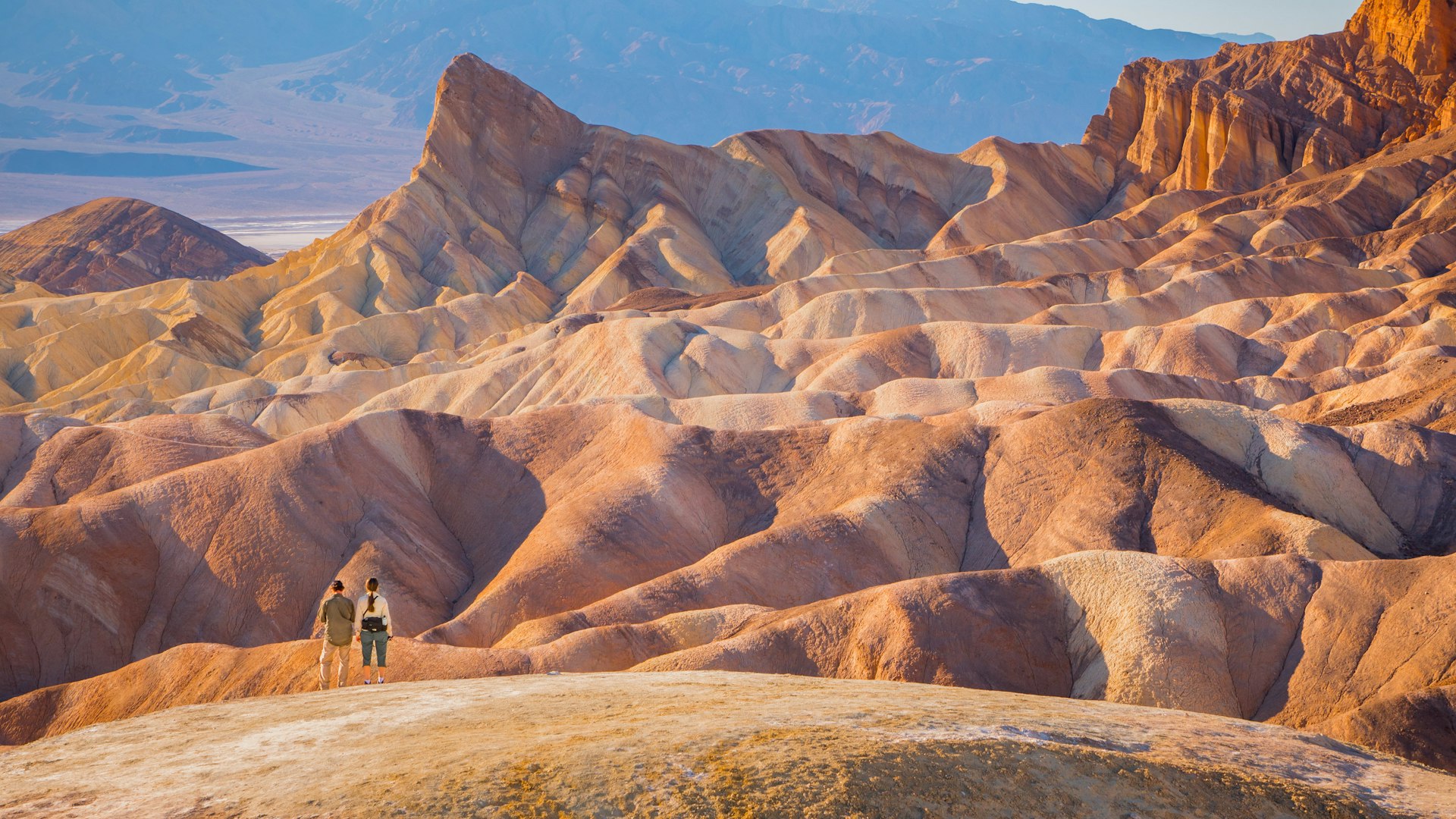 Two people stand amid the rocky, rumpled landscape of Death Valley National Park, looking tiny in comparison the the peaks