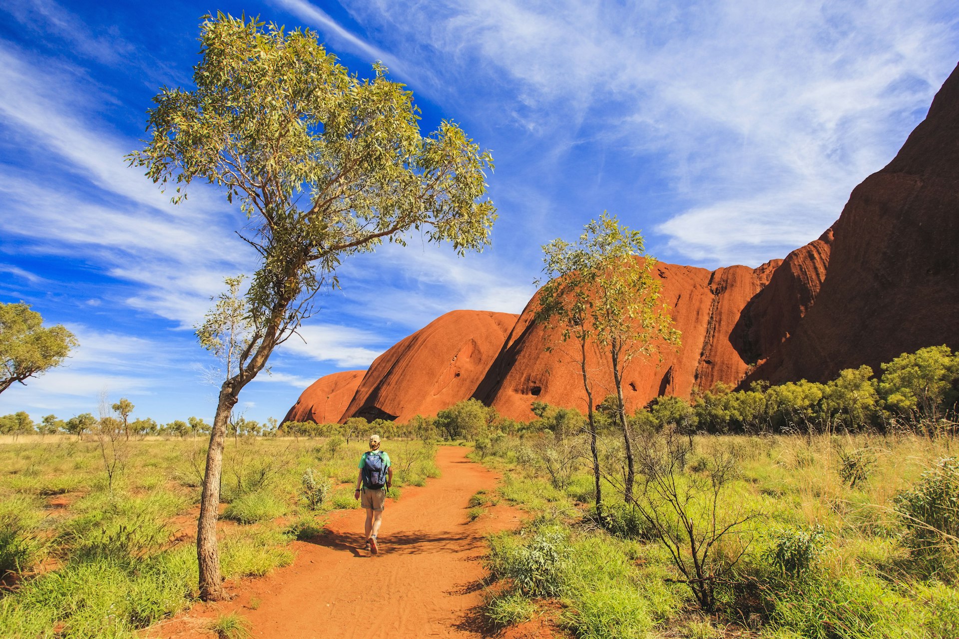 A hiker walks a path around a vast red rock in a sparse landscape