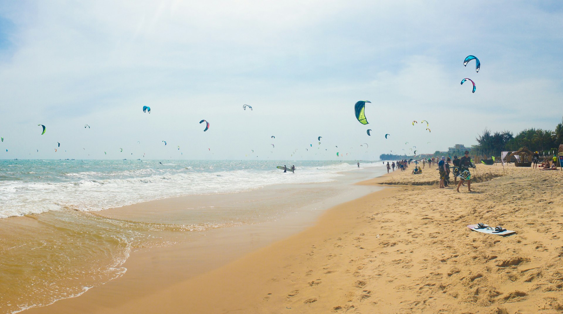 Mui Ne beach in Vietnam. The beach is a long white strip of sand, while the sea is filled with kite surfers, whose colourful kites are visible in the sky above.
