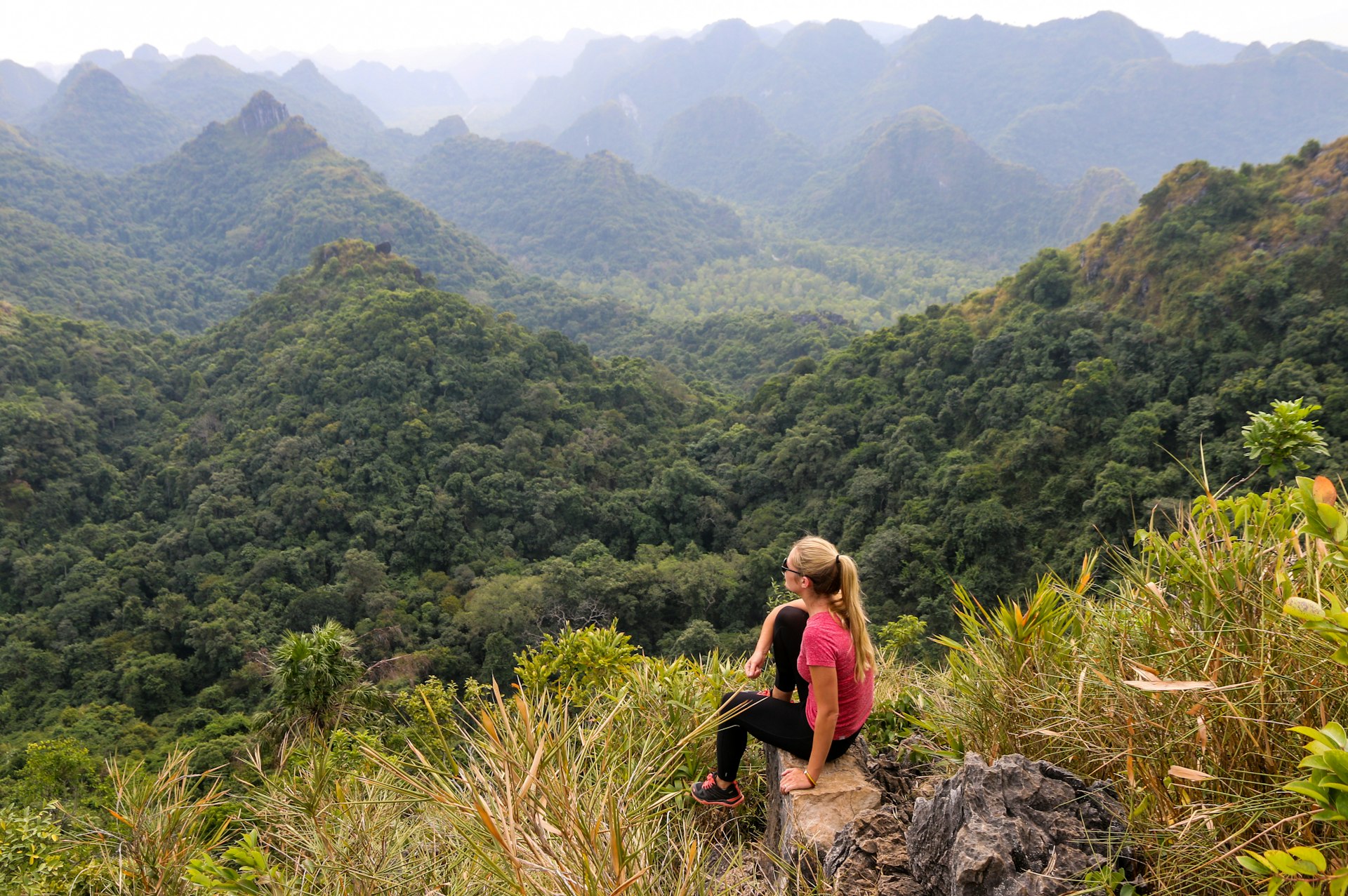 A young woman sitting on a rocky edge and looking at the hills of Cat Ba National Park. The surrounding hills are covered in green forest.
