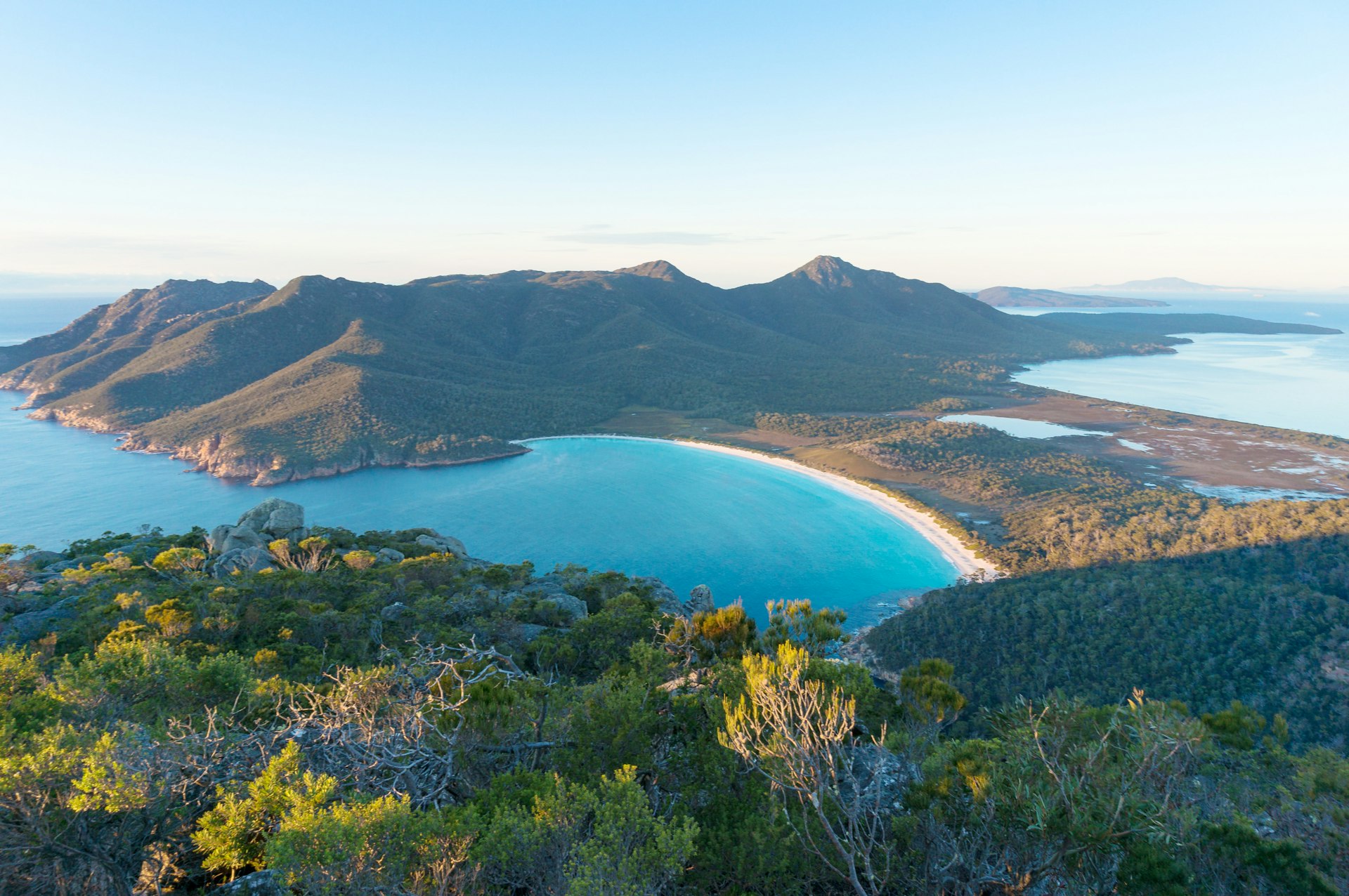 An aerial shot of Wineglass Bay in Tasmania, Australia. The bay's water is turquoise and the arc of the beach is enclosed by greenery.