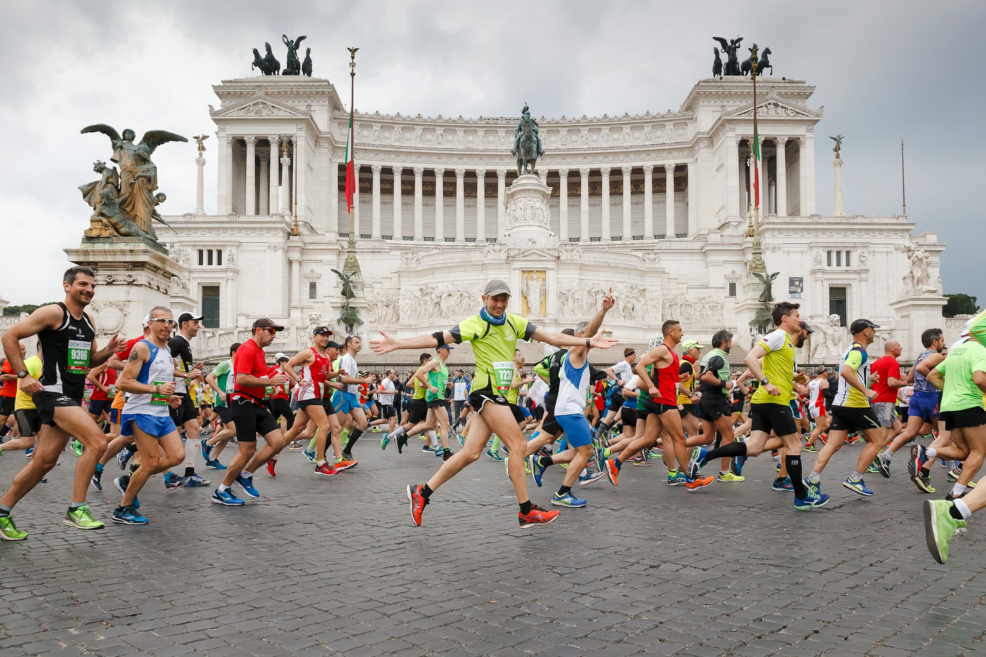 Runners run in the Rome marathon passing a huge white building with columns and several massive brass sculptures