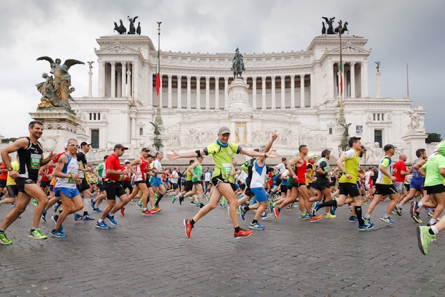 Runners run in the Rome marathon passing a huge white building with columns and several massive brass sculptures