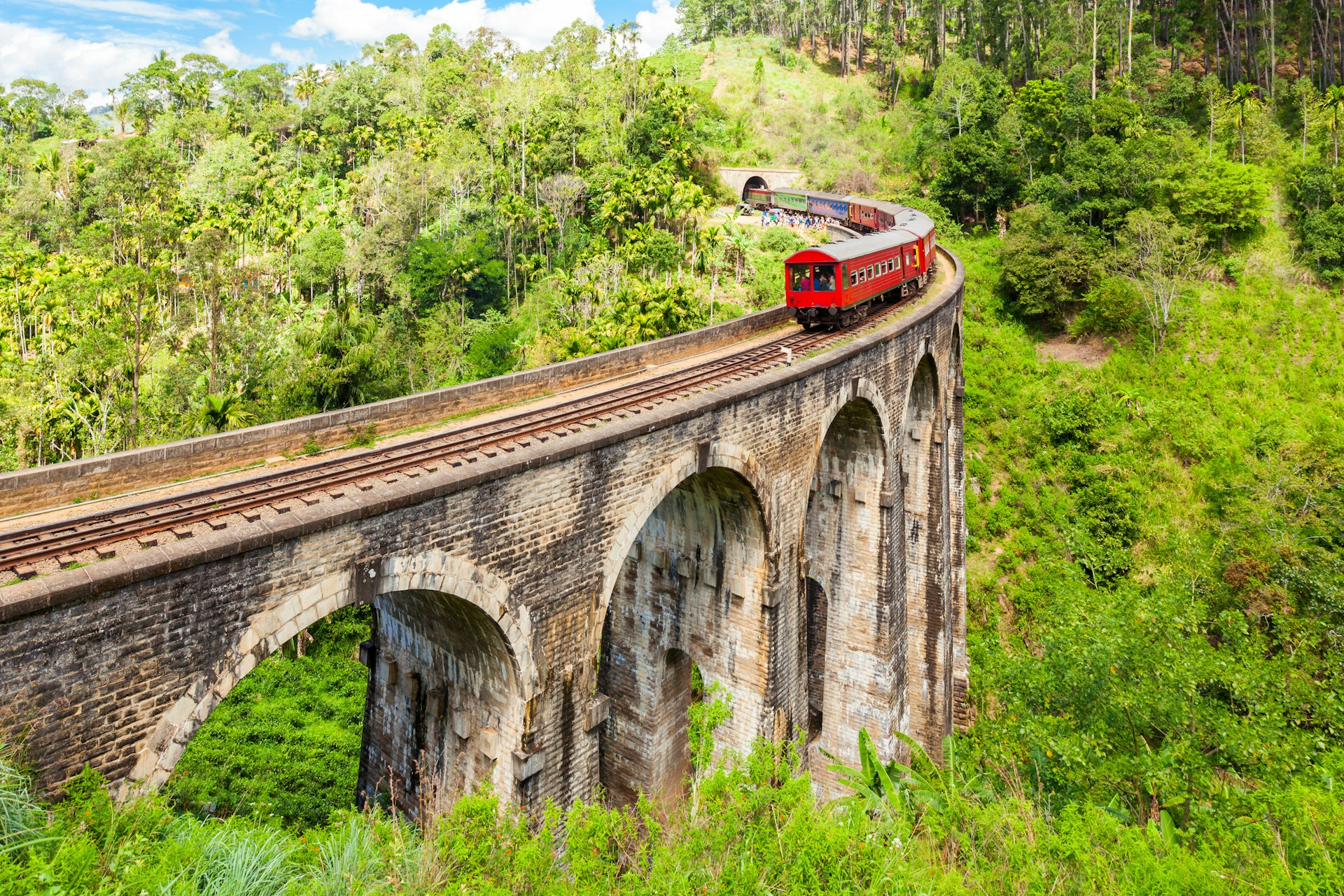 A red train crosses an ornate wooden bridge in the Sri Lankan Hill Country, near the town of Ella. The rolling hills in the background are covered in forest.
