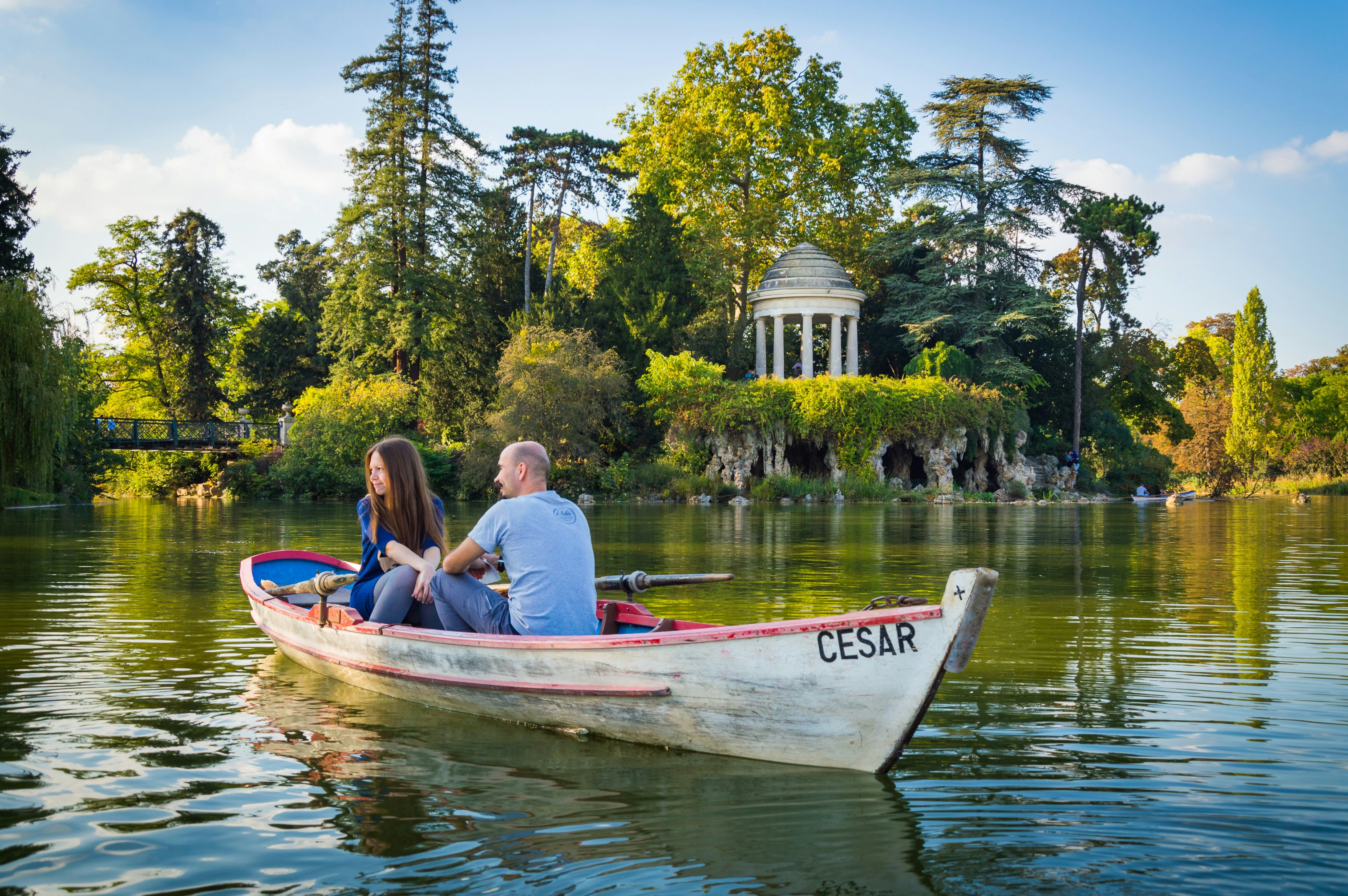 A couple in a row boat passes in front of lush trees.