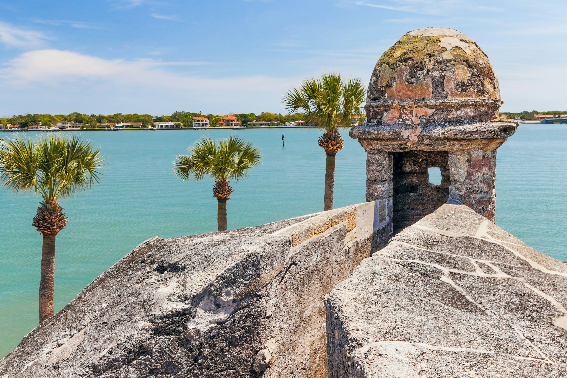 A shot taken from a stone fortress with a rounded sentry turret in the foreground. A palm-tree-lined bay of water stands behind the fort