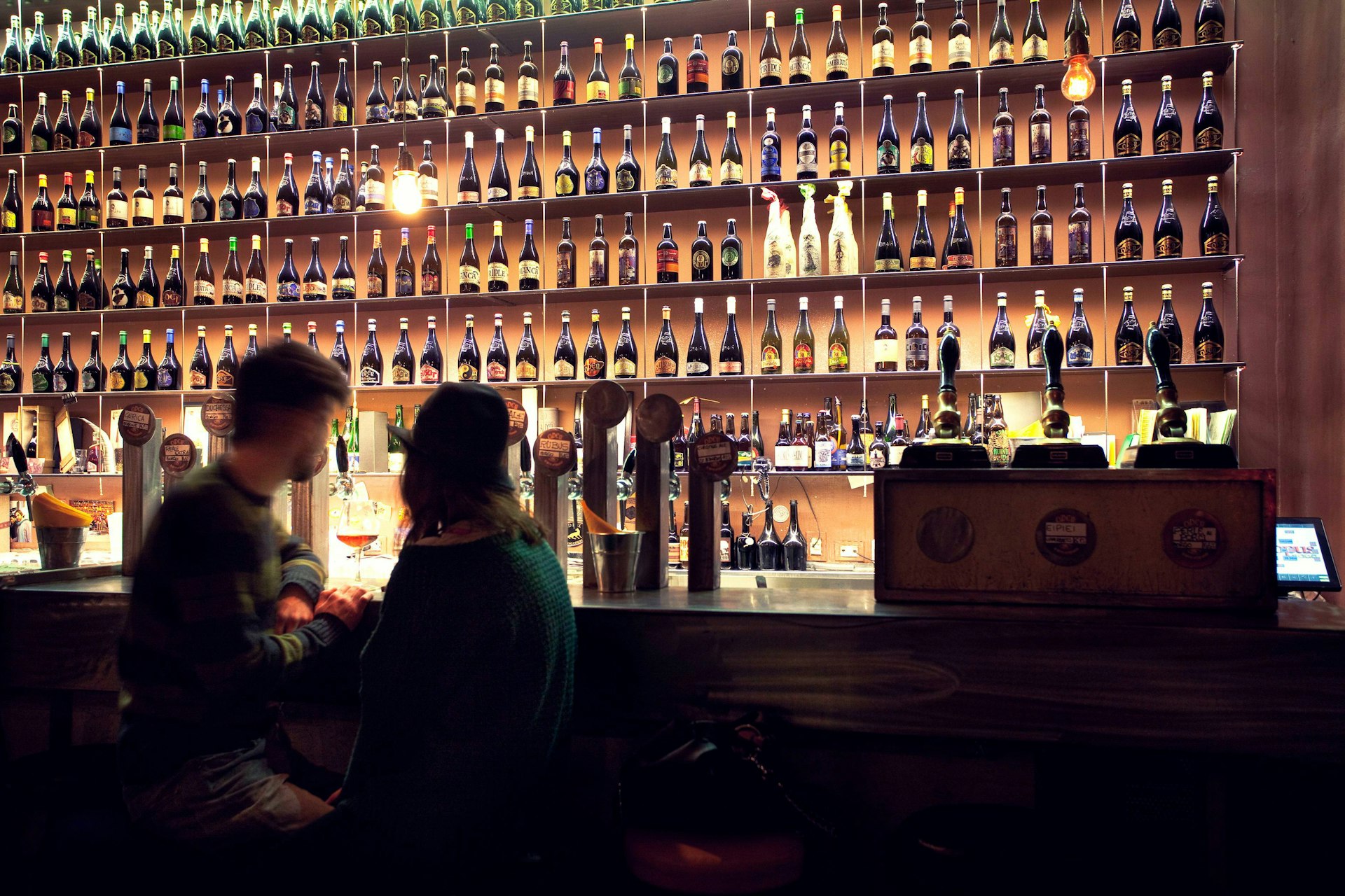 Two people sit at a bar that's backed by hundreds of wine bottles