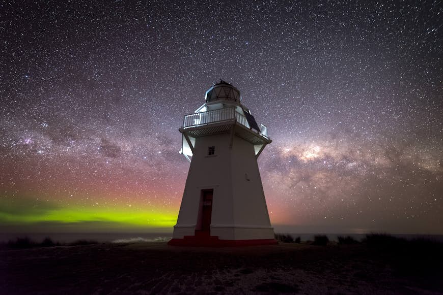 A view of the glowing Southern Lights illuminating the dark night sky behind Waipapa point lighthouse, New Zealand, which stands tall in the foreground.