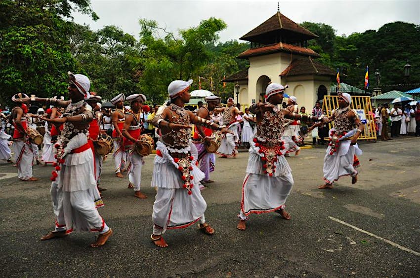 A group of men in traditional clothing dance and play instruments during a religious festival in Kandy, Sri Lanka. The men are all dressed in white and are barefooted.