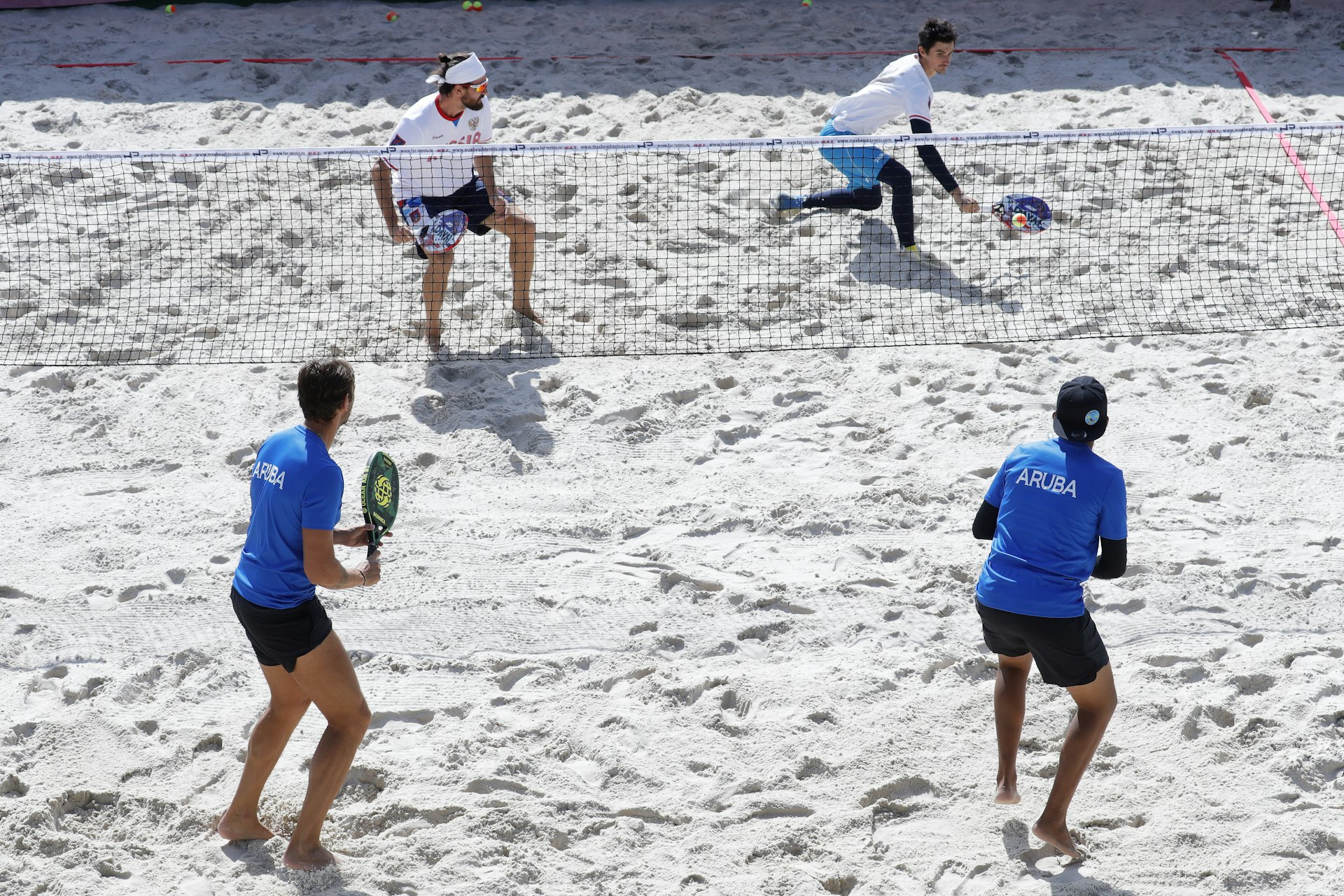 A pair of two-man teams, one wearing blue tops from Aruba and the other wearing white tops from Russia, play tennis on white sand.
