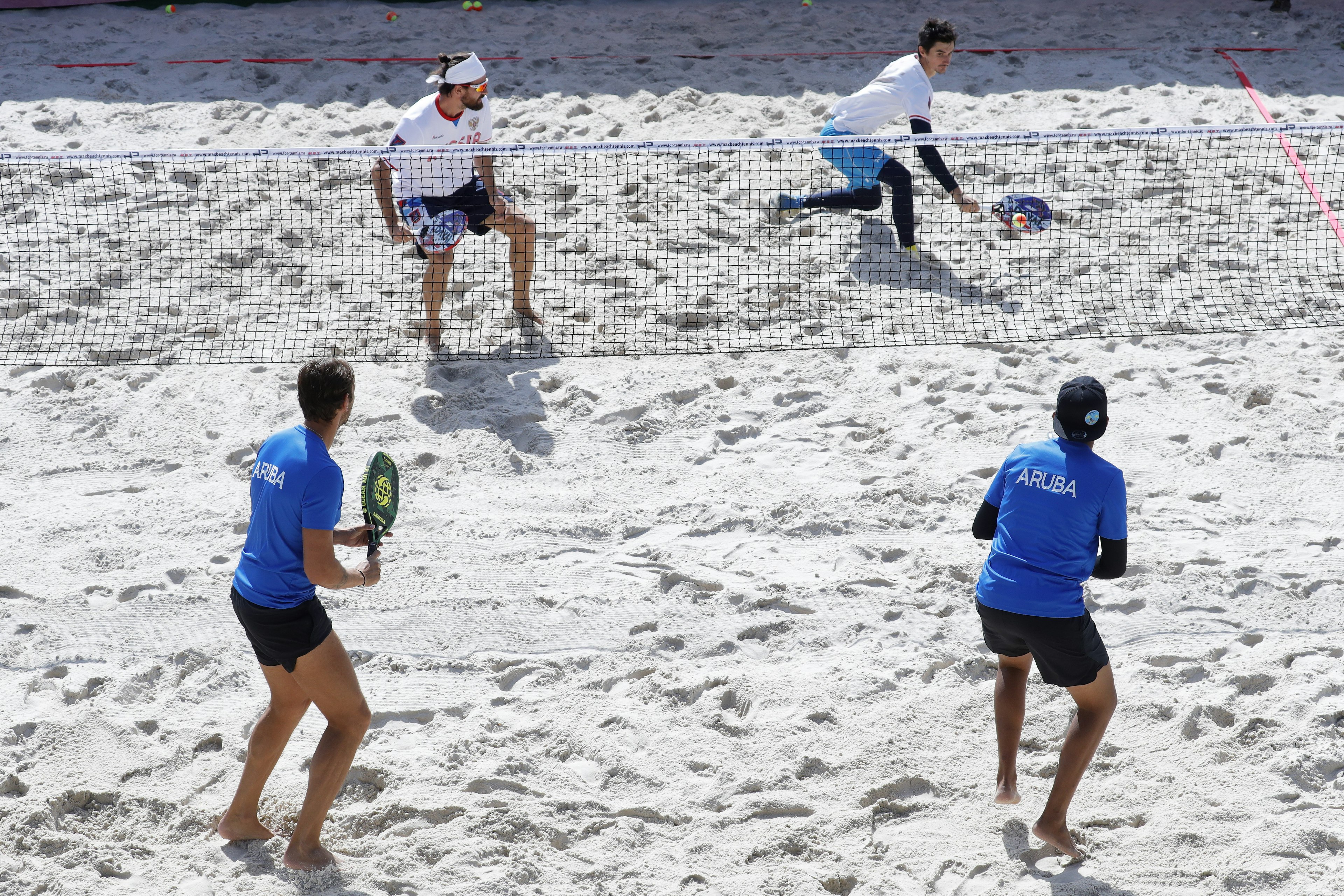 A pair of two-man teams, one wearing blue tops from Aruba and the other wearing white tops from Russia, play tennis on white sand.