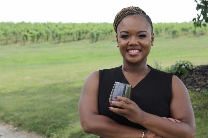 A woman wearing a black dress smiles at the camera while holding a glass of wine. In the background is a grape vineyard. 