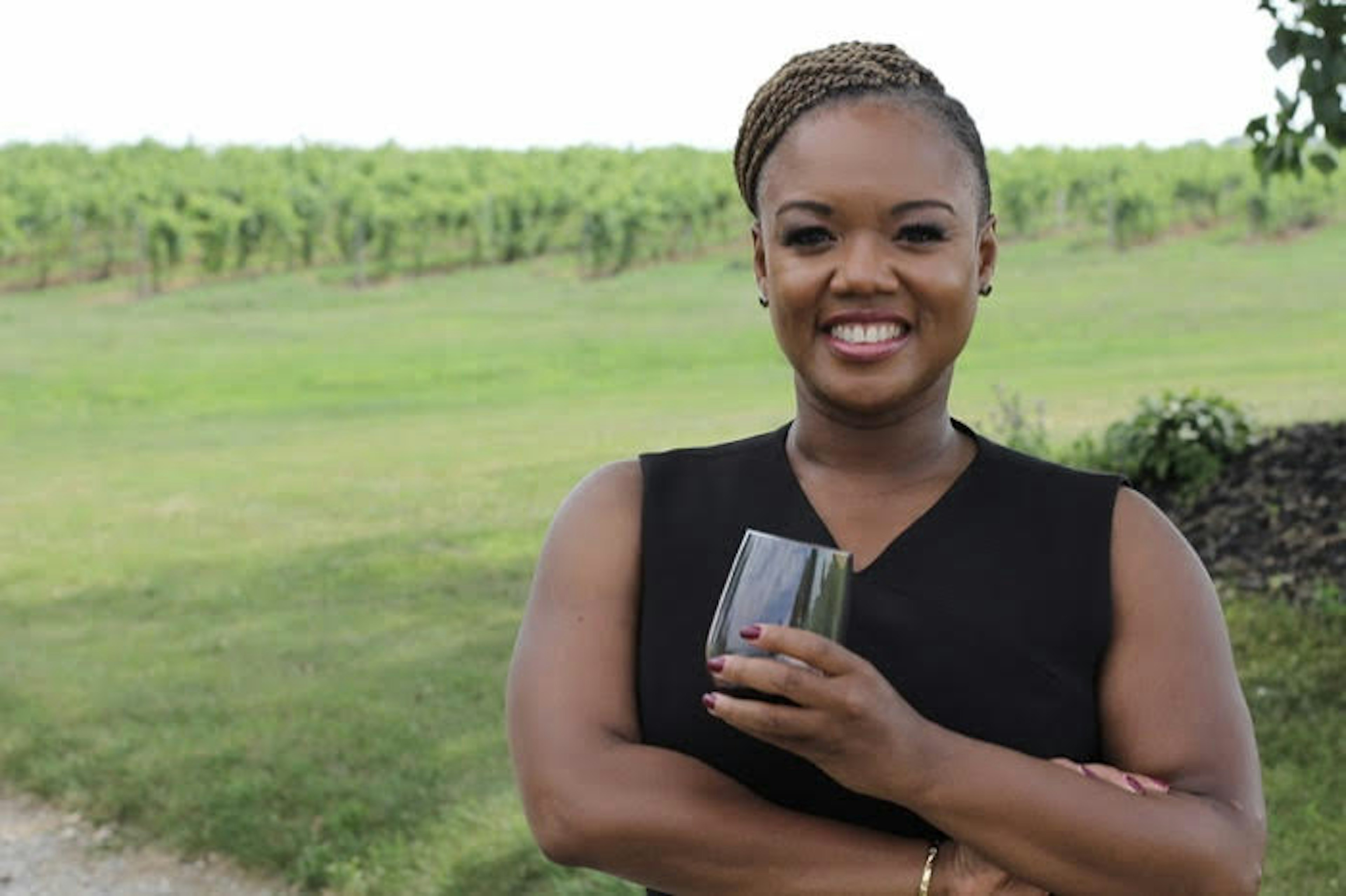 A woman wearing a black dress smiles at the camera while holding a glass of wine. In the background is a grape vineyard.