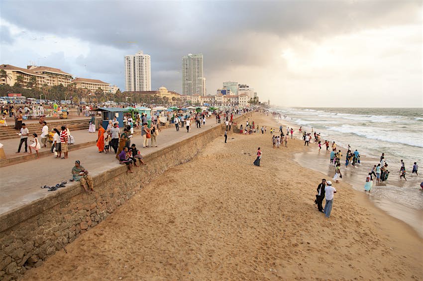 A large crowd walk along a stone promenade and sit on a thin sandy beach on the outskirts of Colombo, Sri Lanka. The park in the background is Galle Face Green.