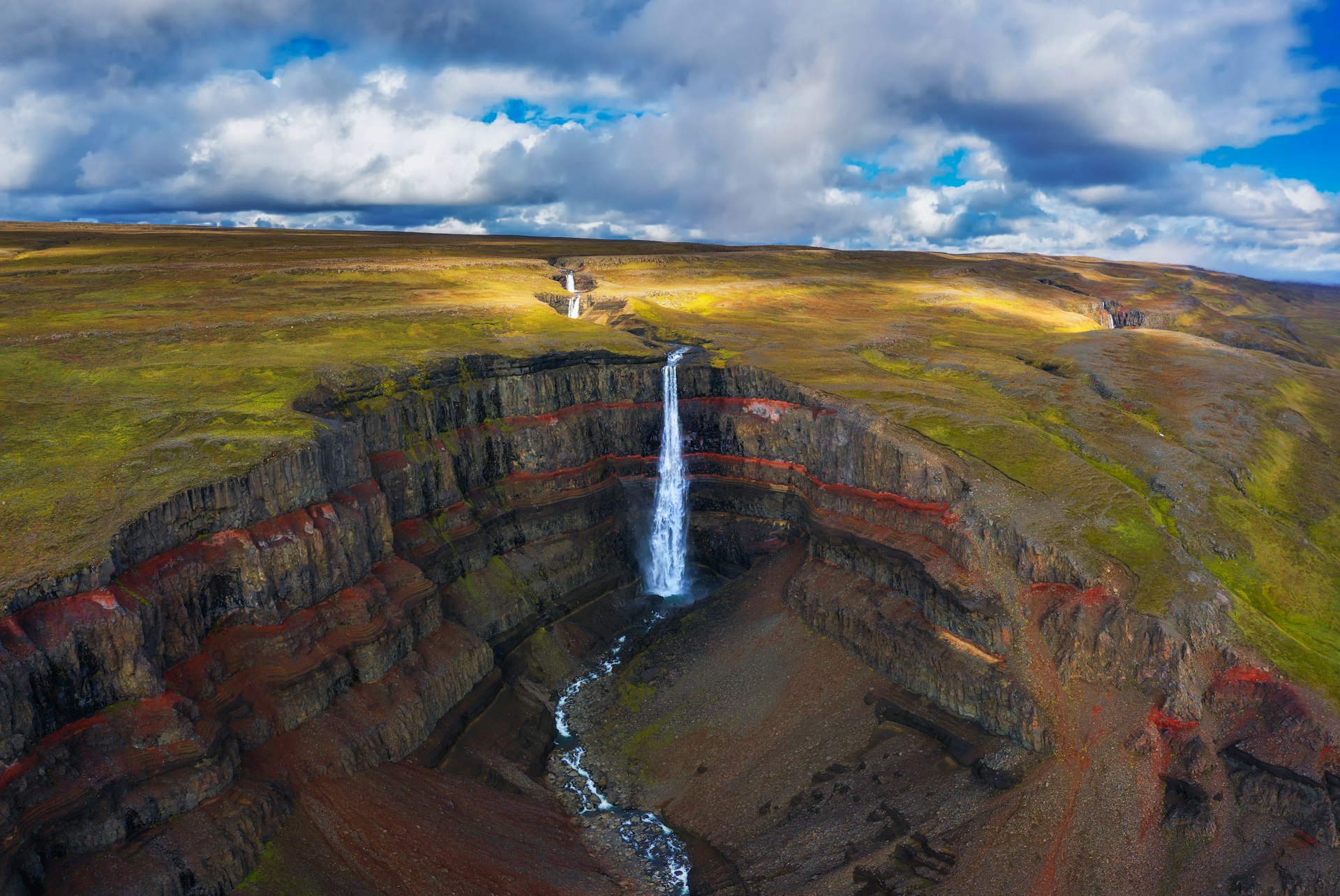 Aerial view of a high waterfall falling down into a crater with red layers of clay between the basaltic layers of rock