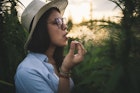 Side view of beautiful woman with hat smoking joint in marijuana plantation at sunset.