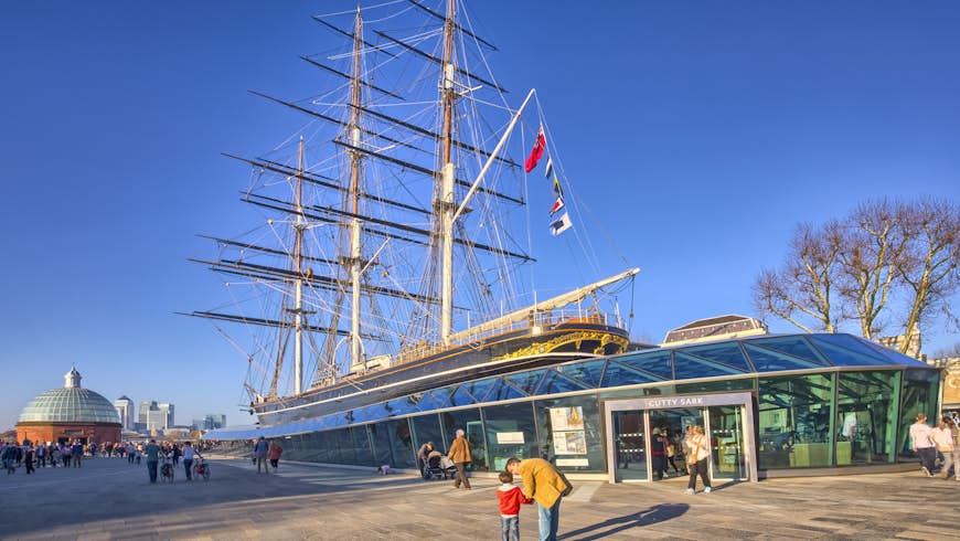 A tea clipper boat on land surrounded by glass to form a museum building. A man kneels down next to a child in the foreground, pointing upwards at the boat's masts.