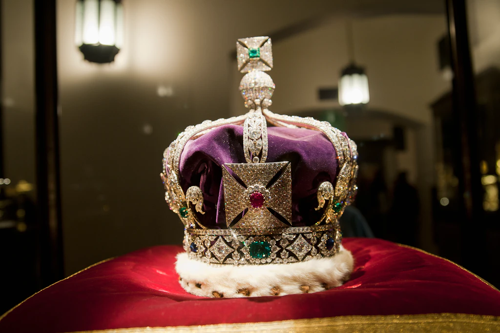 A crown covered in large and smalls jewels sits on a red cushion 