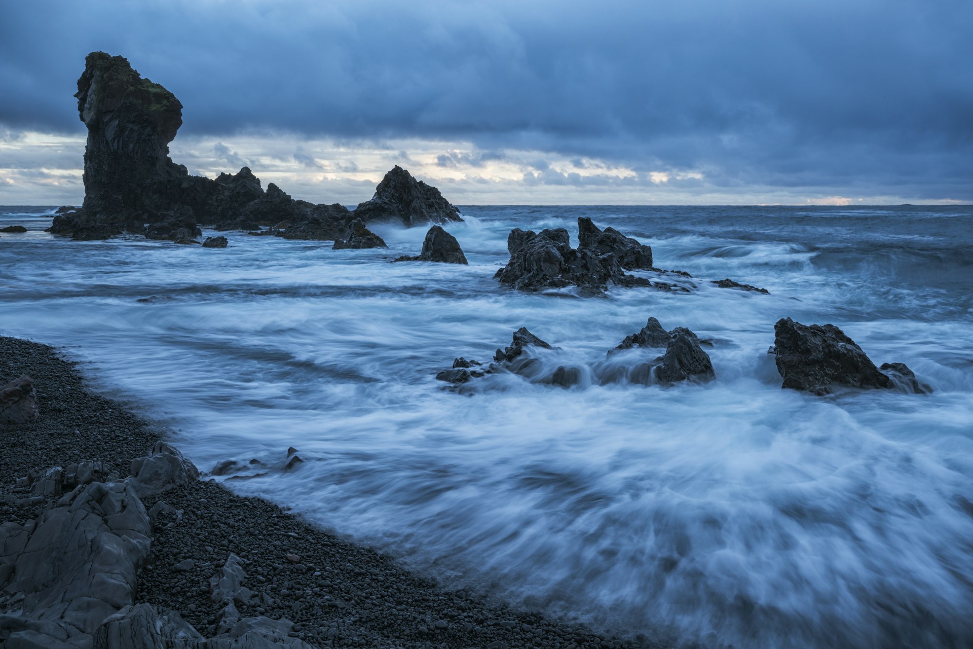 A black sand beach with pounding surf. Several huge rocky stacks stand tall out at sea.