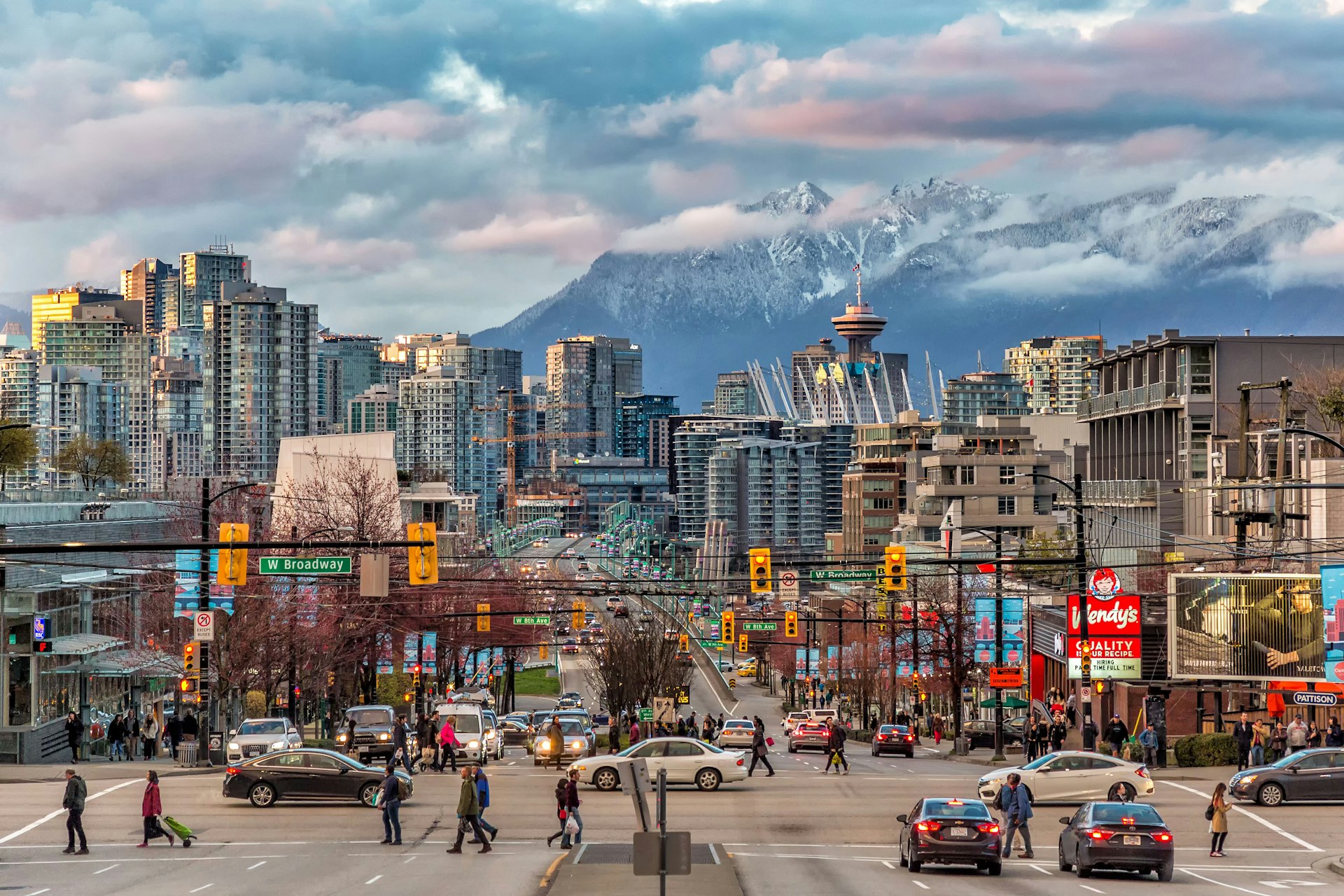 A crossing at Downtown Vancouver where cars wait at the traffic lights and people cross the road during the day with snow-capped mountains across the strait in the background.