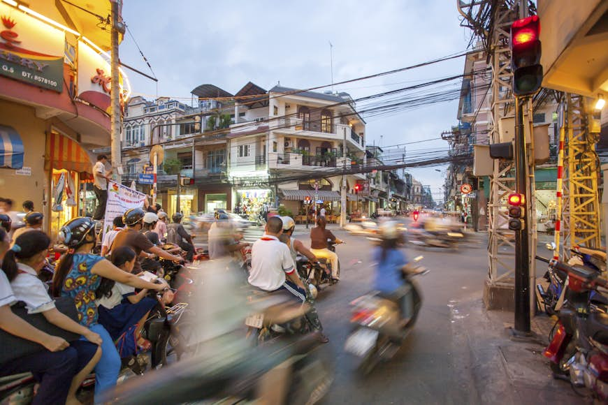 A number of blurred moped cross an intersection in the heart of Ho Chi Minh City. There are many mopeds on the street, making the roads look very busy.