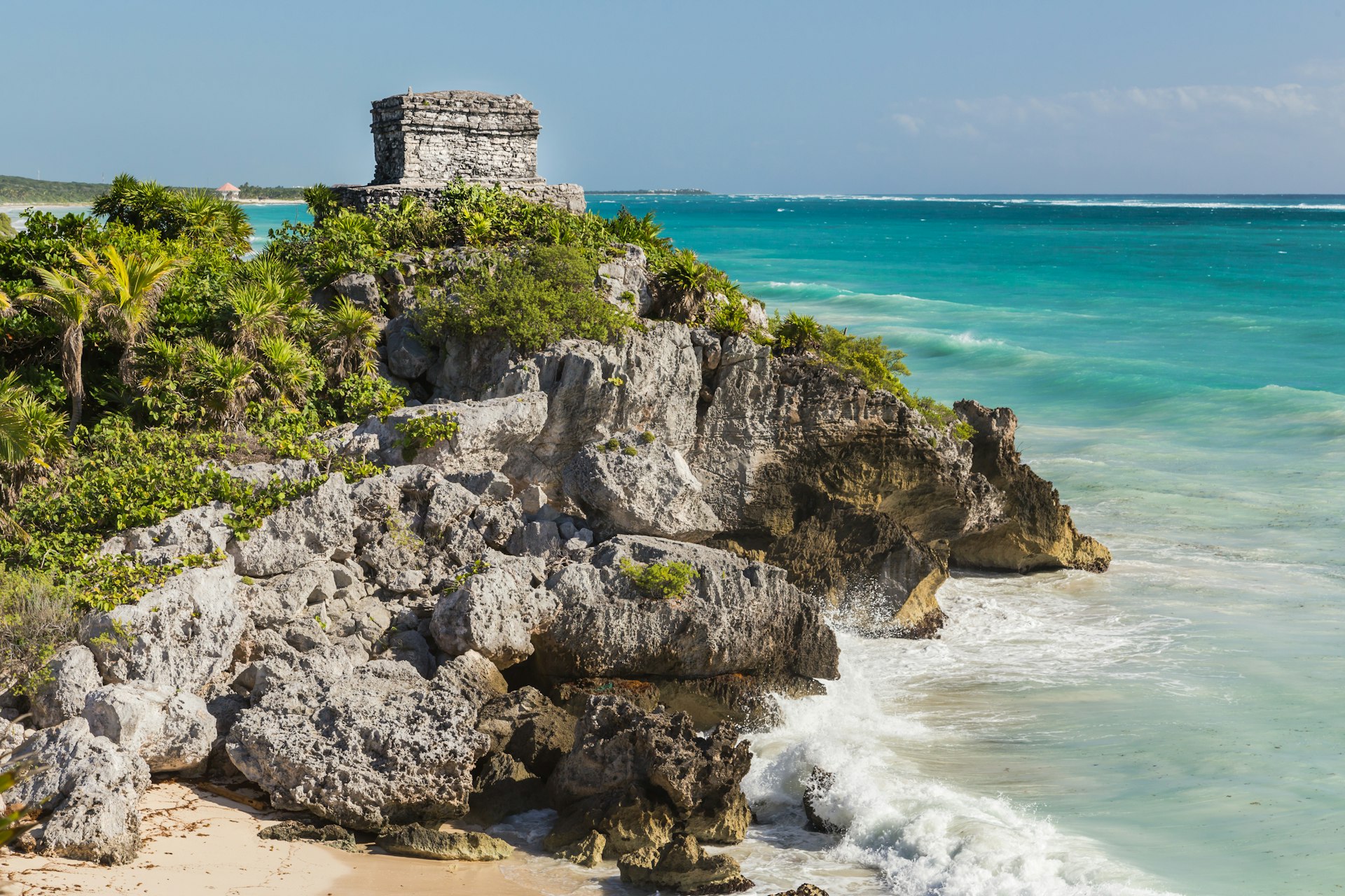 A ruined structure stands at a cliff above a sandy beach surrounded by tropical vegetation