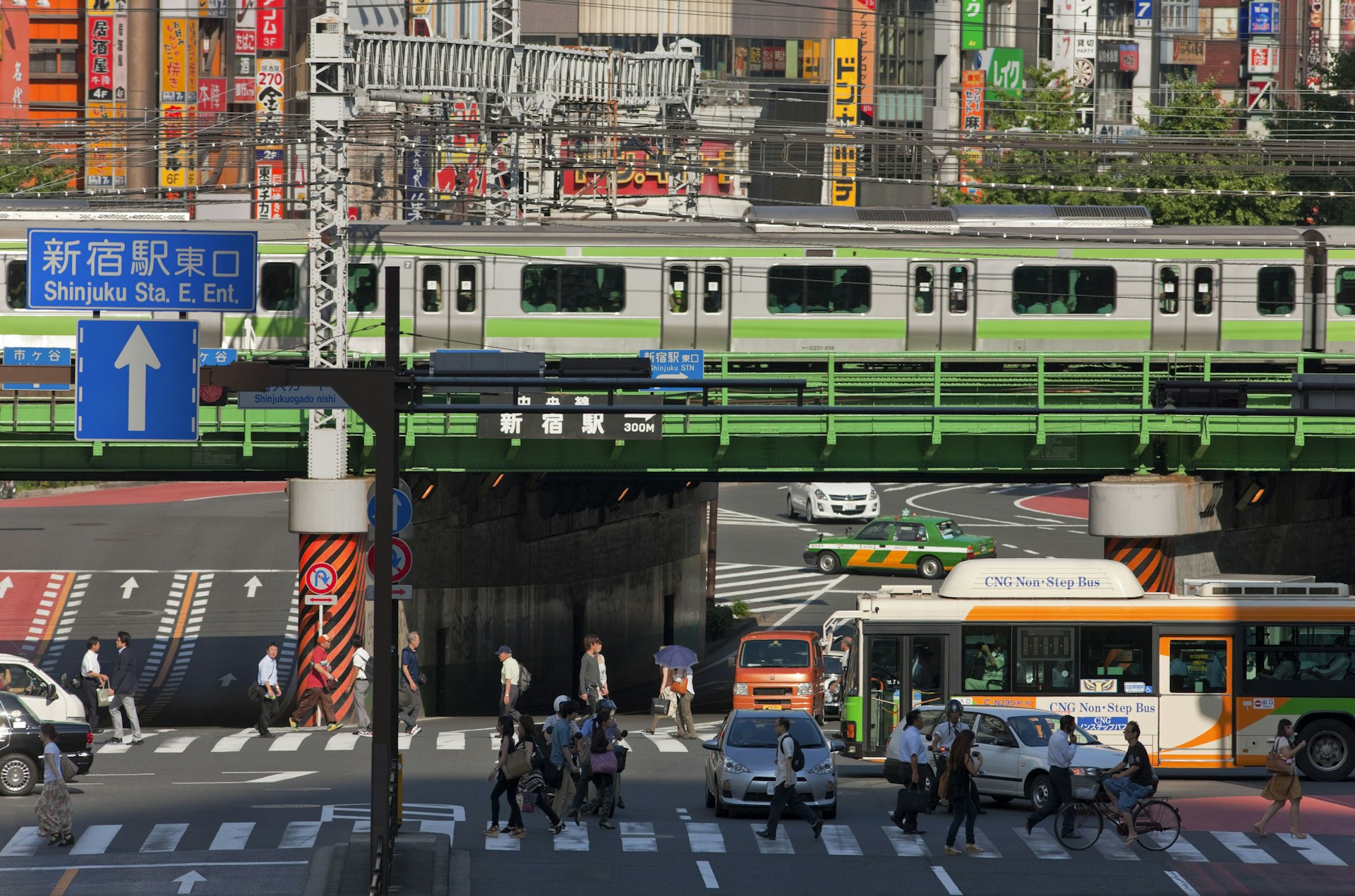A train travels over a bridge above roads with pedestrians, cars and a bus below