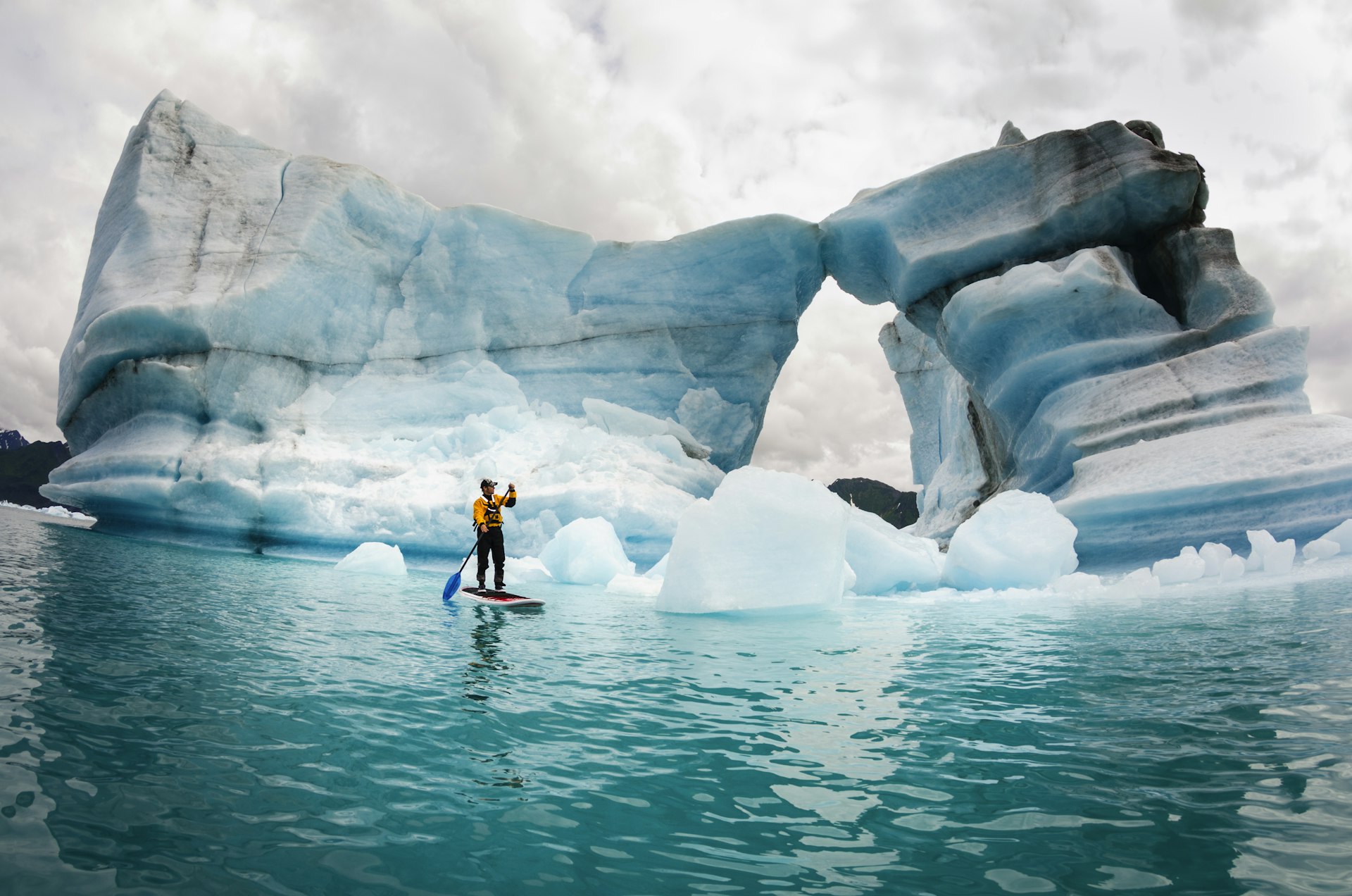 A solo figure on a stand up paddle board (SUP) paddles past icebergs