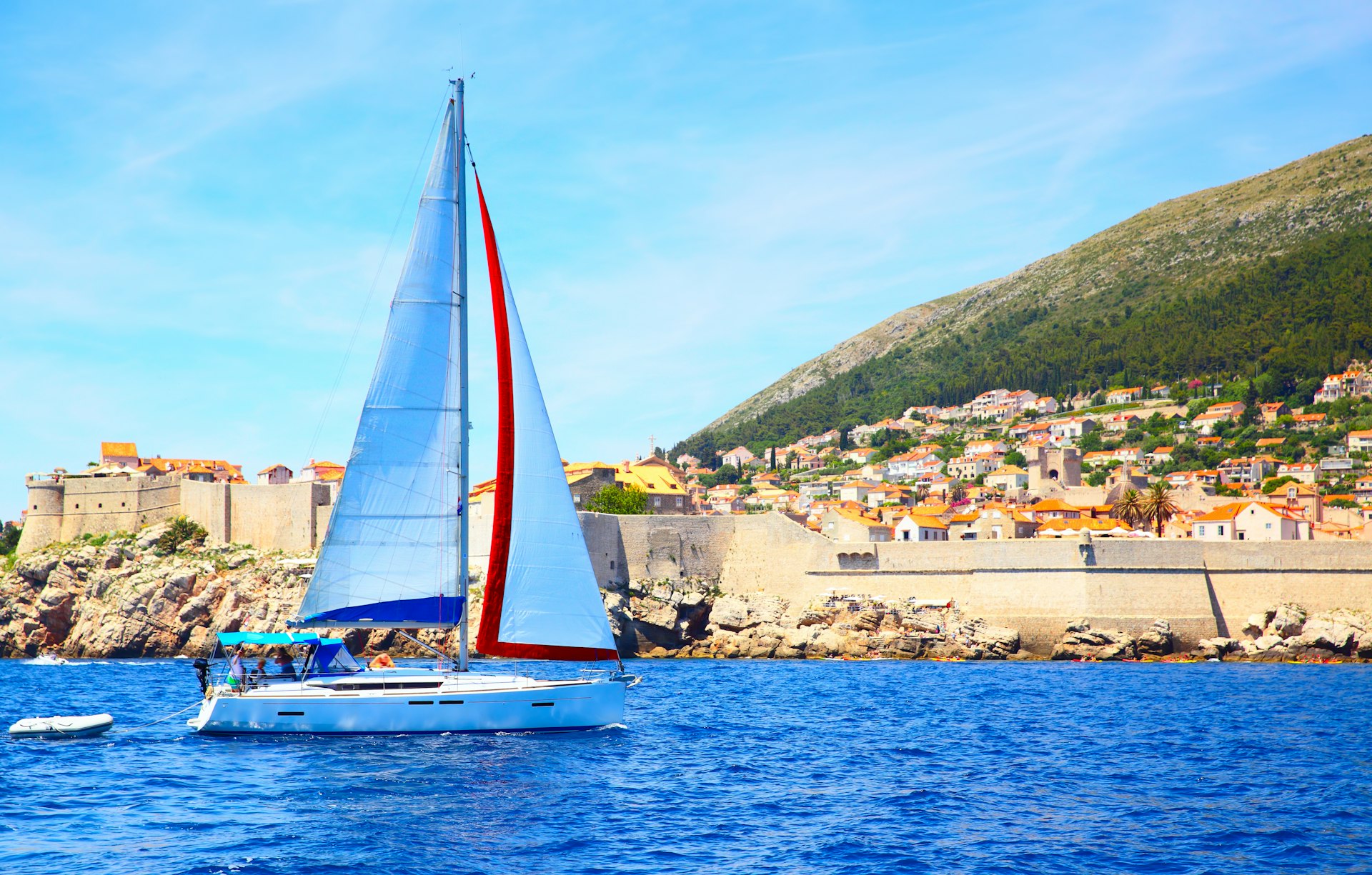 A sail boat sails past Dubrovnik. The small boat has white sails and is surrounded by clear blue ocean.