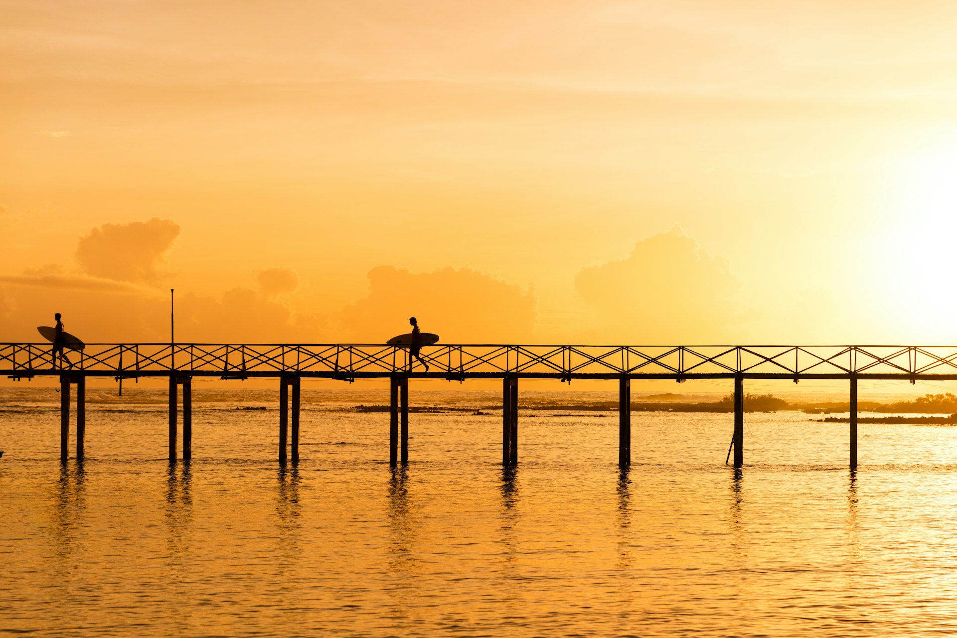 Two people walk across the famous Cloud 9 surf boardwalk in Siargao, the Philippines. The silhouette of the two men is cast against an orange sky with the sun setting.