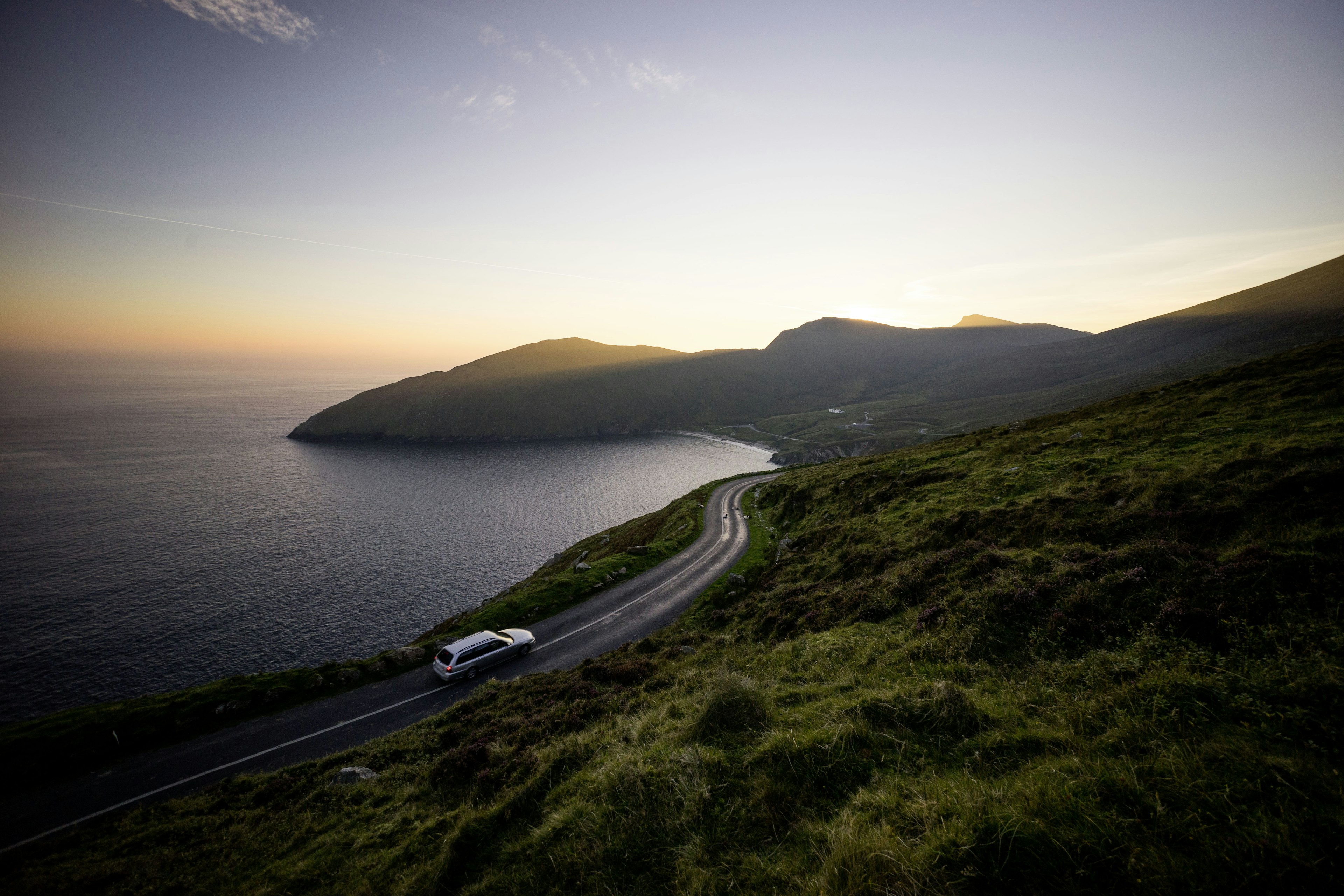 A car drives along a coastal road as the sun sets