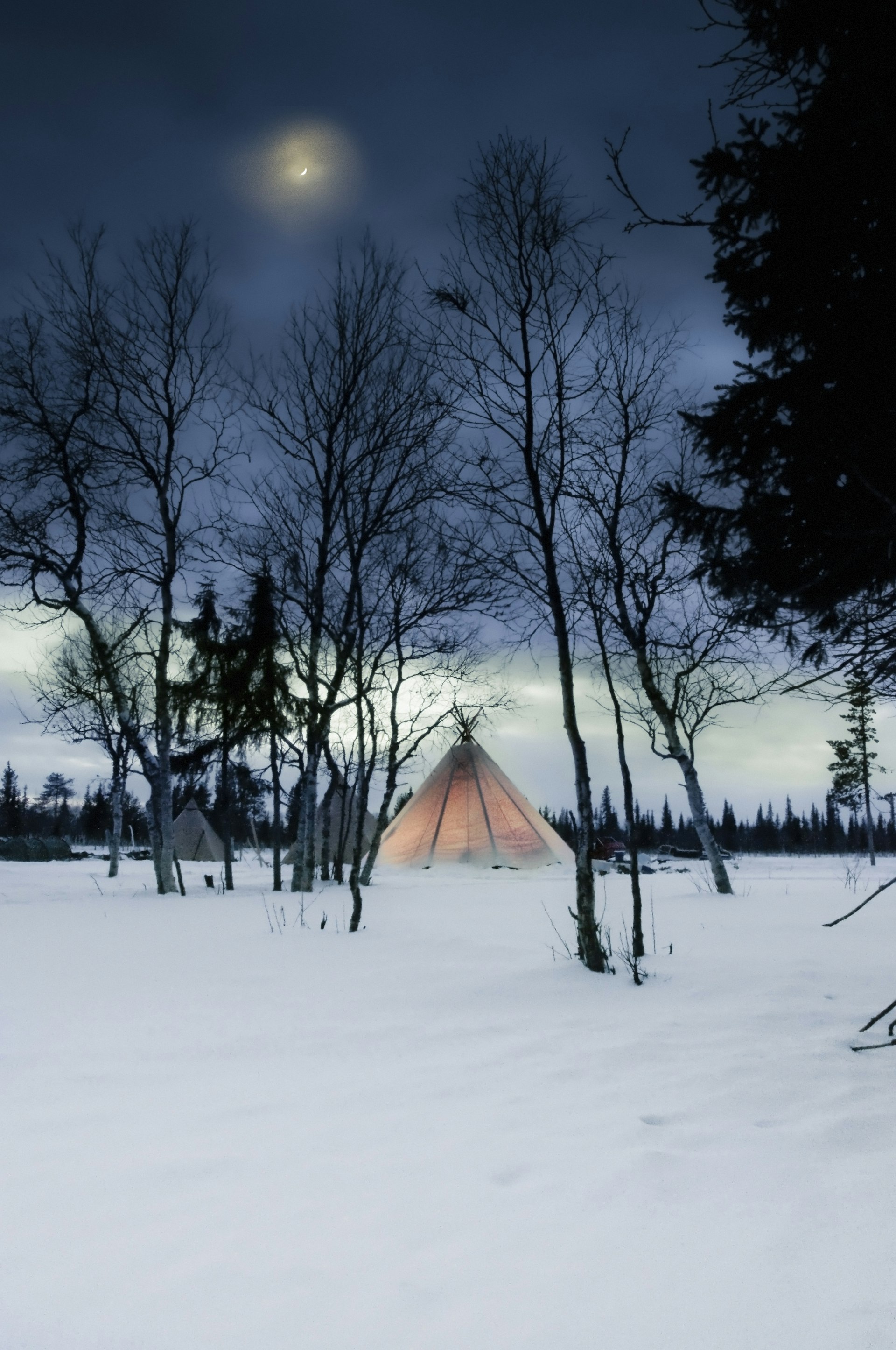 A traditional Sami tent glows with light from inside. It is in the middle of a snow-covered forest.