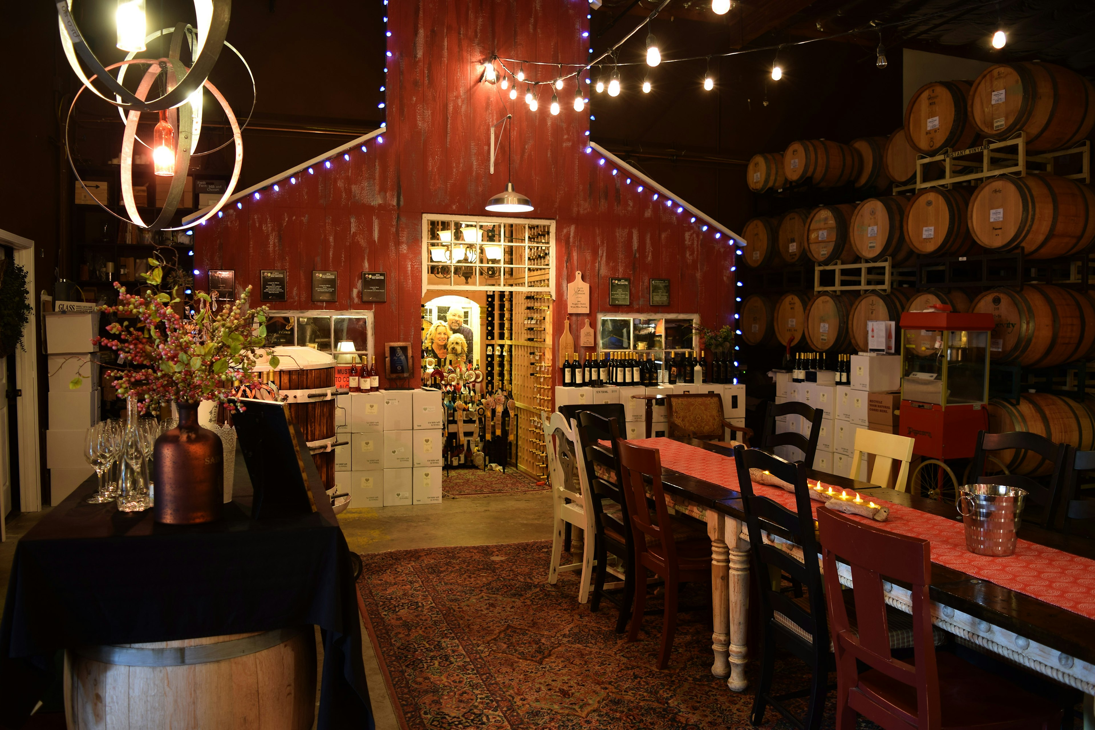 Interior shot of the Longevity Tasting Room. There is a large structure outlined with lights. There are wine barrels stacked on the right and a long wooden table in the middle. You can see a man, a woman and a dog peeking through the door in the back of the room.