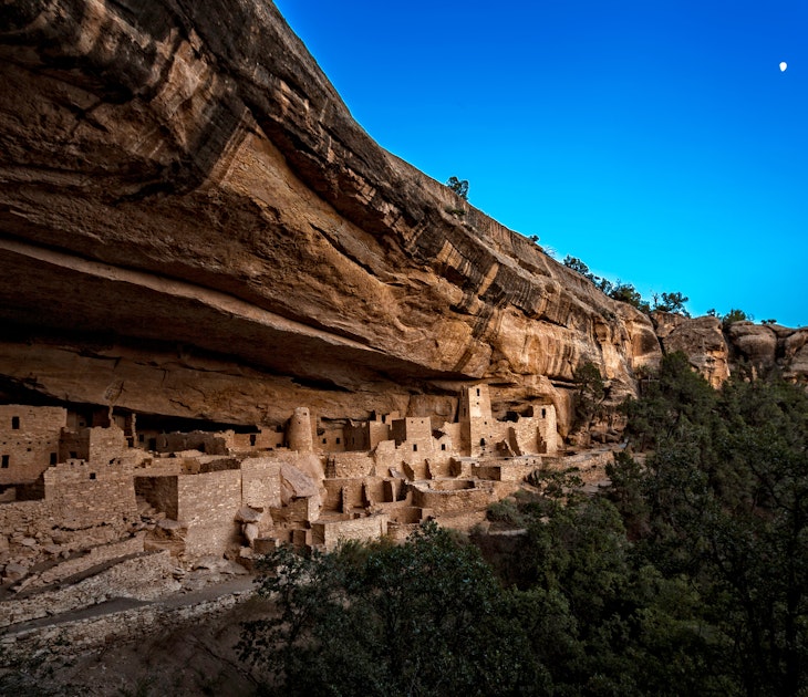 A twilight image with a half moon in a clear blue sky over the Cliff Palace ruins in Mesa Verde National Park, with the pink-beige brick structures in the light and everything else in shadow