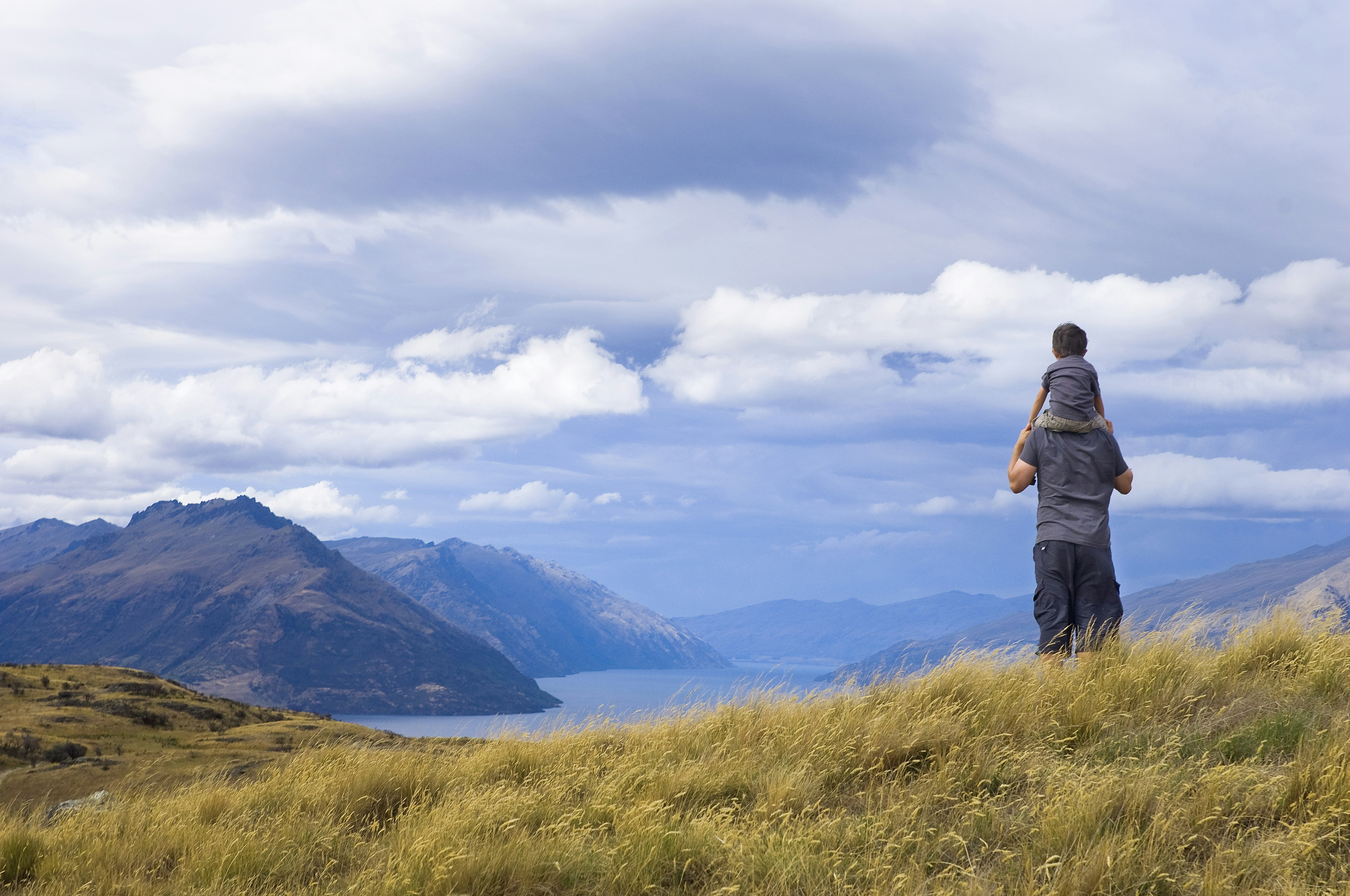 A child sits on the shoulders of his father whilst looking out to the sea and mountains of New Zealand