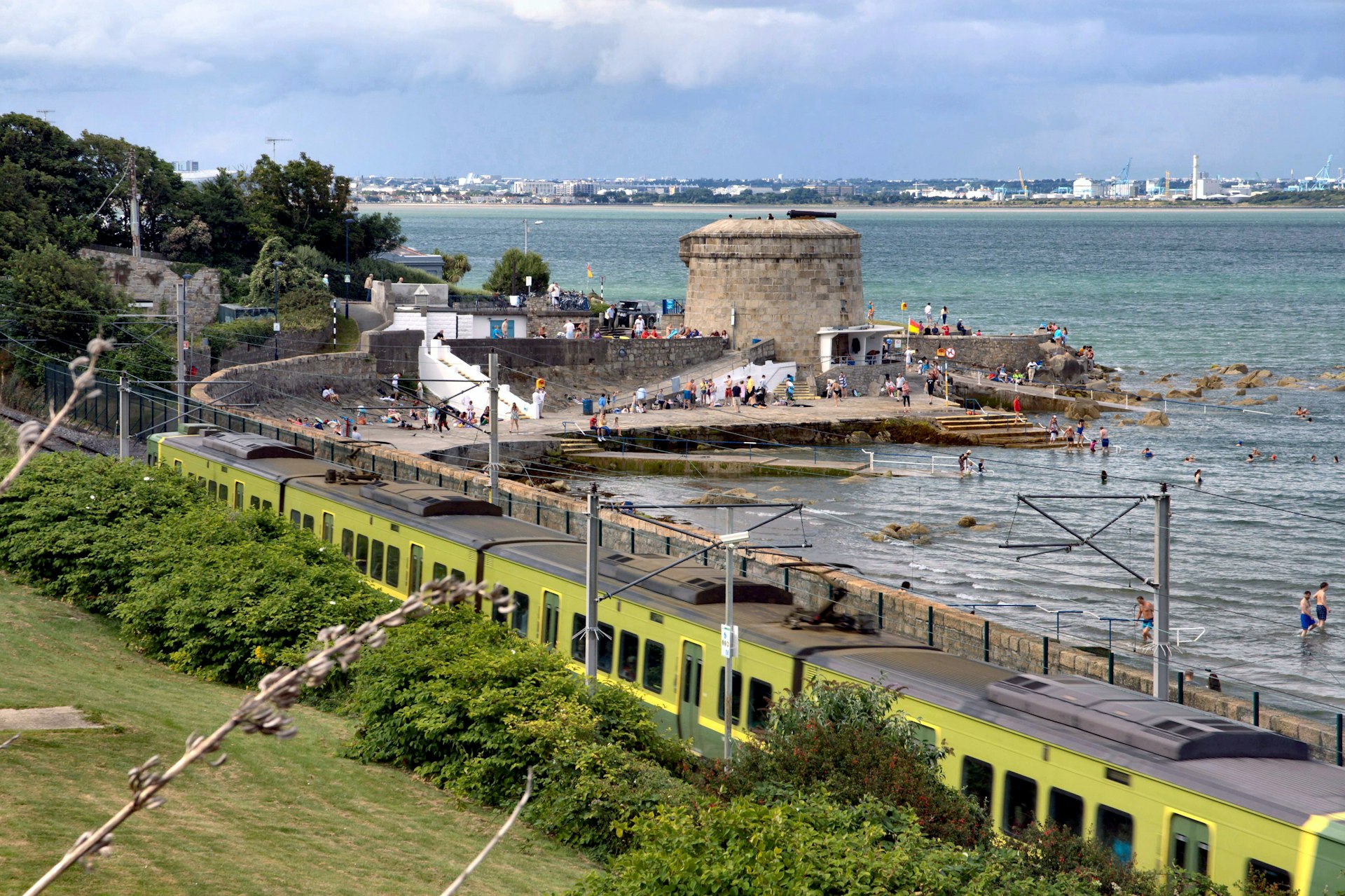 A DART train passes by a busy Seapoint Beach on a cloudy day in Dublin