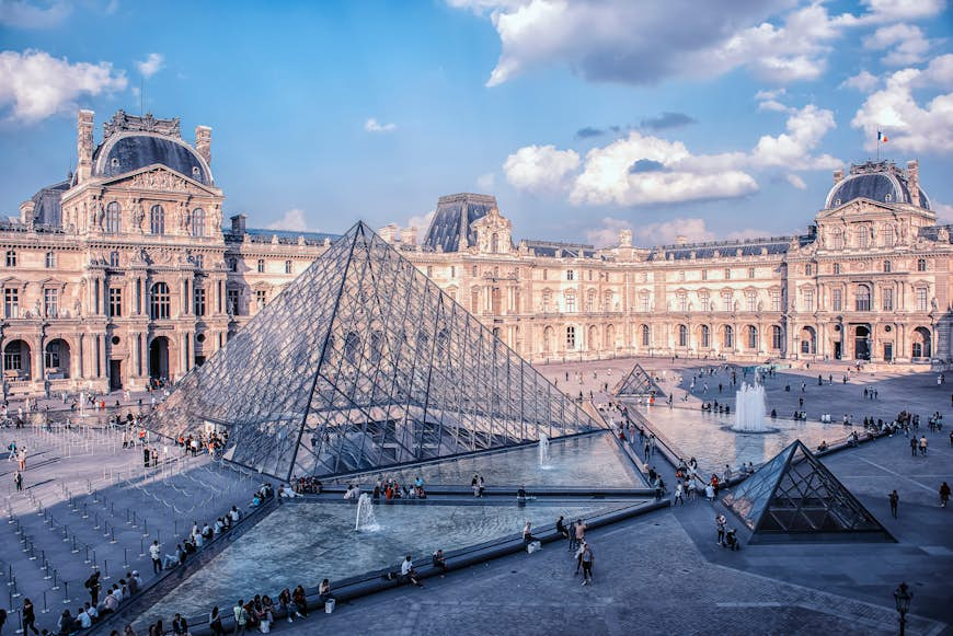 People walk around outside the Louvre museum in daytime. 