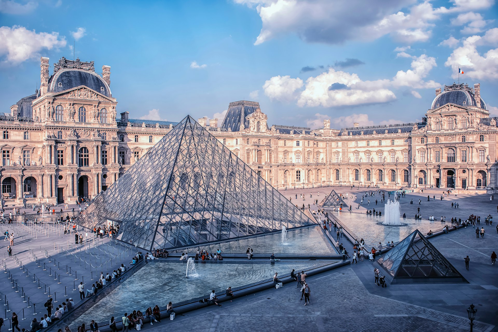 People walk around outside the Louvre museum in daytime. 